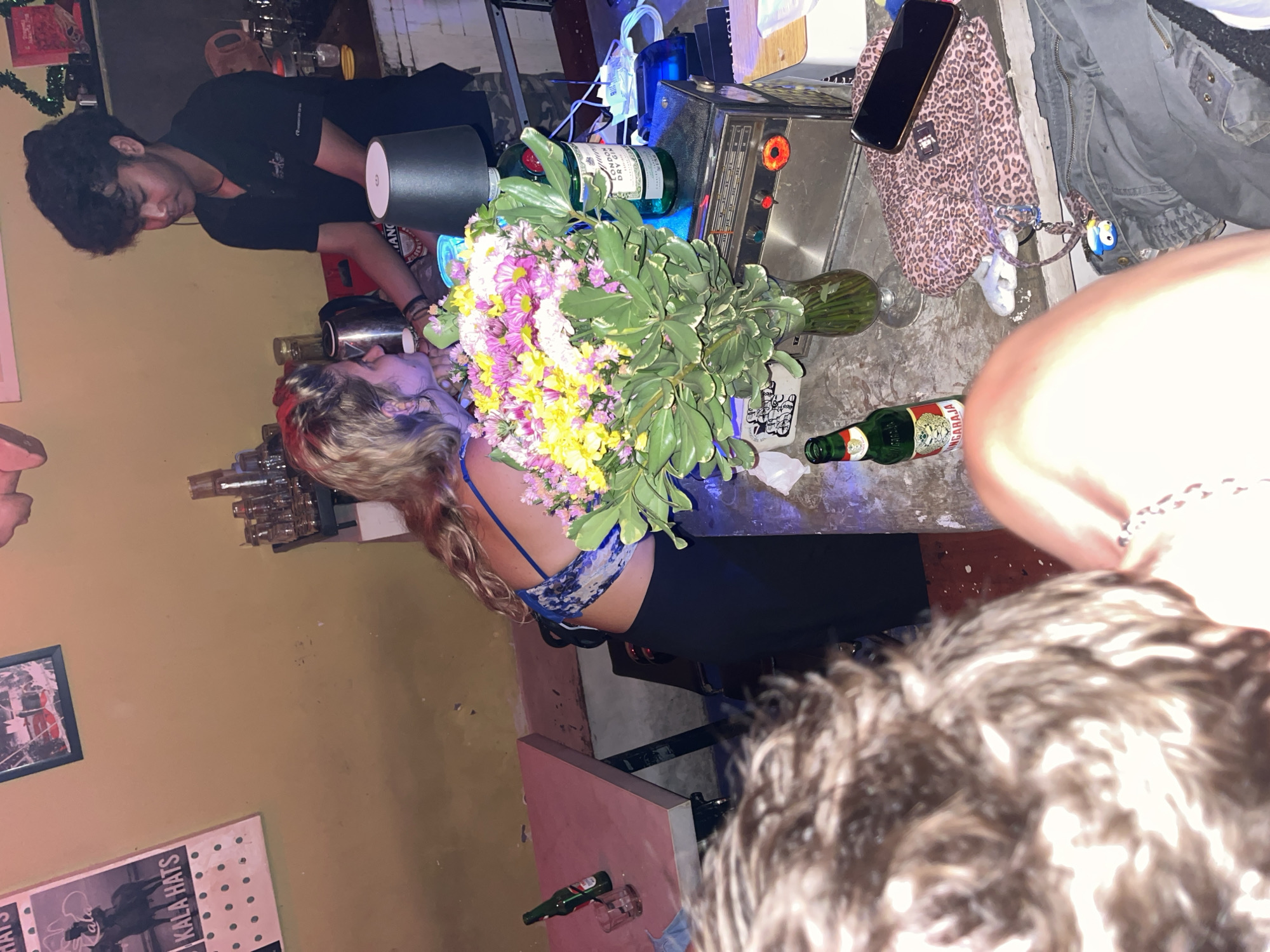 This image captures a lively, casual scene inside a bar or pub, likely in North Kuta, Indonesia, given the provided context. The overall lighting is dim, suggesting it is evening or nighttime.
The main subjects include a male bartender and two female patrons. The bartender, with dark curly hair and wearing a black short-sleeved shirt, is positioned behind a bar counter, attending to something. A woman with blonde, wavy hair, wearing a blue patterned bralette or top and dark trousers, is leaning over the bar counter, looking down or interacting with the bartender. Her face is partially obscured by a large, colorful bouquet of flowers. The head of another person with curly dark hair is visible in the foreground, suggesting a first-person perspective or a close-up shot from another patron's view.
The bar counter is cluttered with various objects. A prominent bouquet of yellow, pink, and white flowers with green foliage sits on the counter. Beside it, a bottle of "LONDON DRY GIN" is visible. Other items include glasses, a shaker, and what appears to be a vintage radio or sound system. On the floor or a lower shelf by the bar, a leopard print bag with a mobile phone on it, a grey jacket, and a keyring with a blue and white character are seen. A green beer bottle with "PABARRIJA" visible on the label lies on its side. In the background, a yellowish-beige wall displays framed pictures and posters. One poster clearly shows "KALA HATS". The scene also features some industrial-style lighting and visible cables, adding to the informal ambiance.