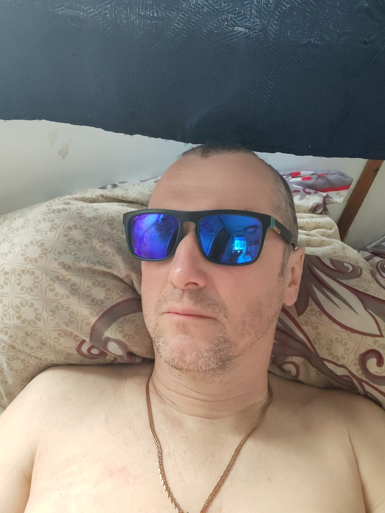 A man is lying on a pillow. He is wearing sunglasses and a gold chain. The environment is a room with a dark fabric hanging above him. The city and location are unknown.