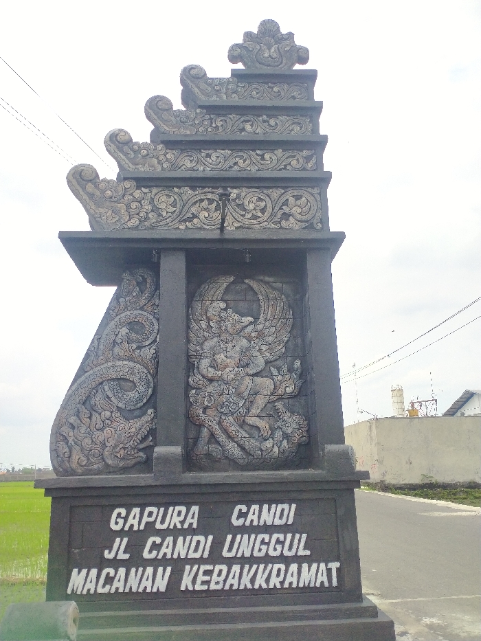 A large, ornate stone gateway, known as a "gapura," stands prominently in the center of the frame. The gate is dark gray with lighter, intricately carved stone details, featuring multiple tiered levels at the top adorned with floral and swirling motifs. Below the upper tiers, on the front face of the gate, there is a large relief carving of a winged, human-like figure, likely Garuda, grasping a serpentine creature or naga. To the left of this central carving, on the side of the gate, another relief depicts a coiled dragon-like figure with scales and a detailed head.
The gate is situated next to a paved road that stretches into the distance on the right side of the image. To the left of the gate, a vibrant green rice paddy field is visible, suggesting an agricultural landscape. In the background, beyond the road and field, are several buildings, including what appears to be industrial structures or tall tanks, and utility poles with overhead wires. The sky is overcast and bright, indicating daytime without direct sunlight.
At the base of the gate, white capital letters against a dark background display text in Indonesian. The text reads:
"GAPURA CANDI"
"JL CANDI UNGGUL"
"MACANAN KEBAKKRAMAT"
This signage identifies the structure as a temple gate and indicates its location on Jalan Candi Unggul in Macanan, within the Kecamatan Kebakkramat area of Indonesia.