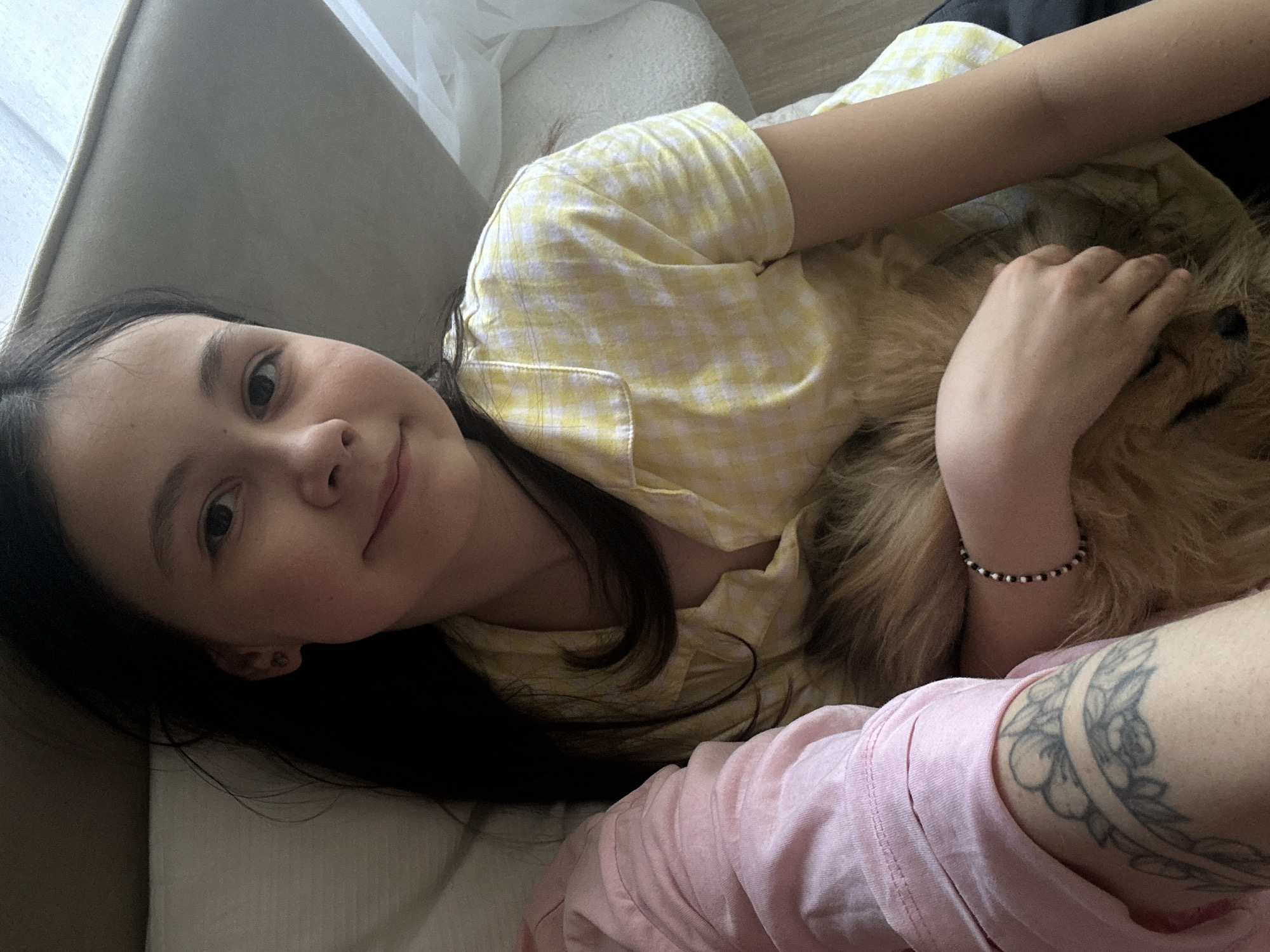A girl with long dark hair, wearing a yellow and white checkered shirt, is lying down. She is looking at the camera and smiling. She is hugging a brown dog. A person wearing a pink long-sleeve shirt with a floral tattoo on their arm is next to her. They are on a light-colored couch. The setting is indoors. The city is not possible to determine from this image.
