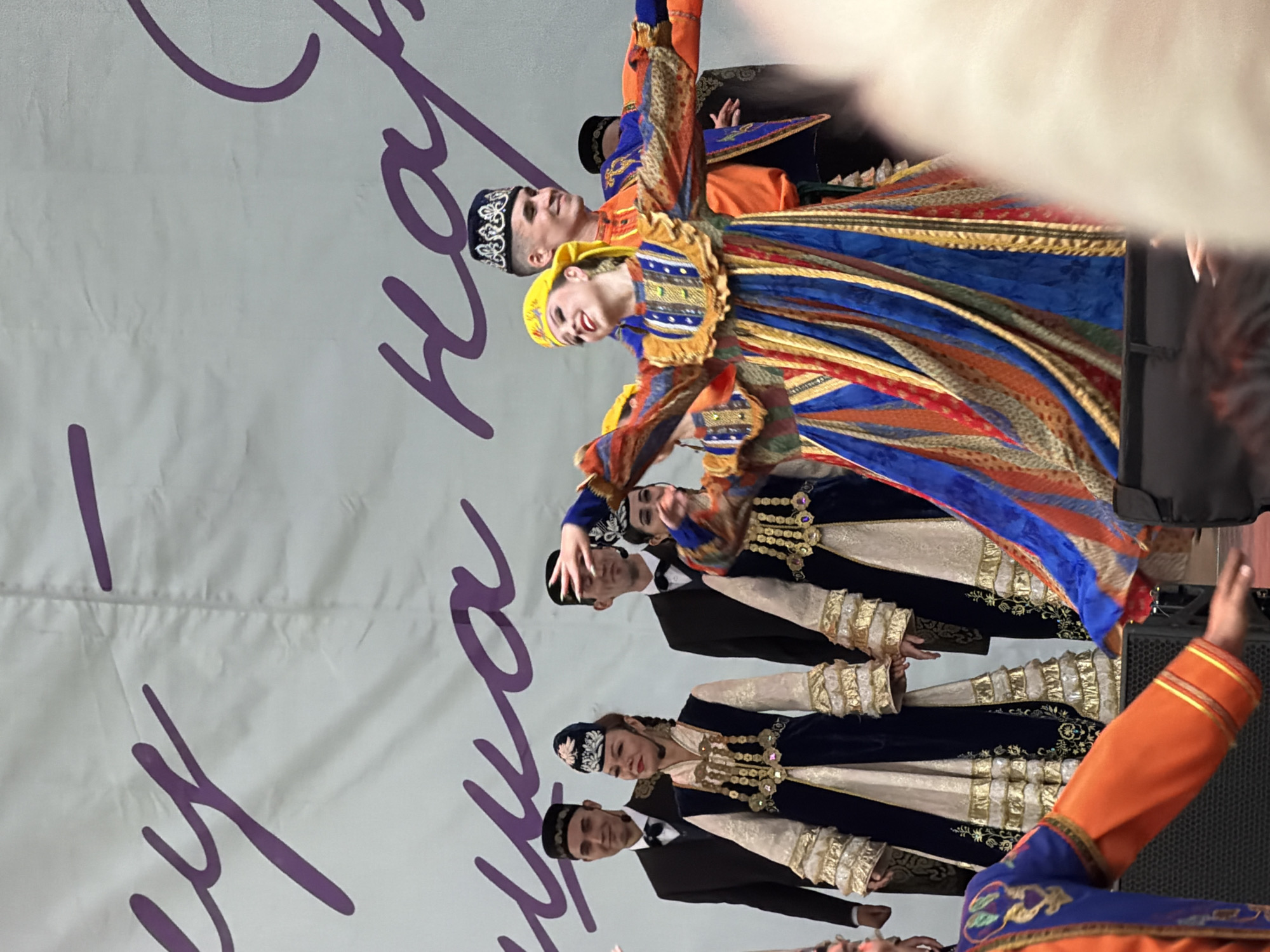 This media file captures a performance by a dance group in Kazan, Russia. The subjects are a group of people, likely dancers, dressed in vibrant, traditional attire. Their costumes are richly decorated with intricate patterns and embellishments, featuring a mix of blues, oranges, reds, and gold. Some dancers wear distinctive embroidered hats. They appear to be in motion, suggesting a dance performance or rehearsal.
The background consists of a large, light-colored fabric, possibly a backdrop or curtain, with large purple lettering in a script font. The lighting suggests an indoor event, likely a performance hall or stage. The overall mood seems energetic and celebratory, judging by the colorful costumes and the dynamic poses of the dancers. The image is oriented vertically.