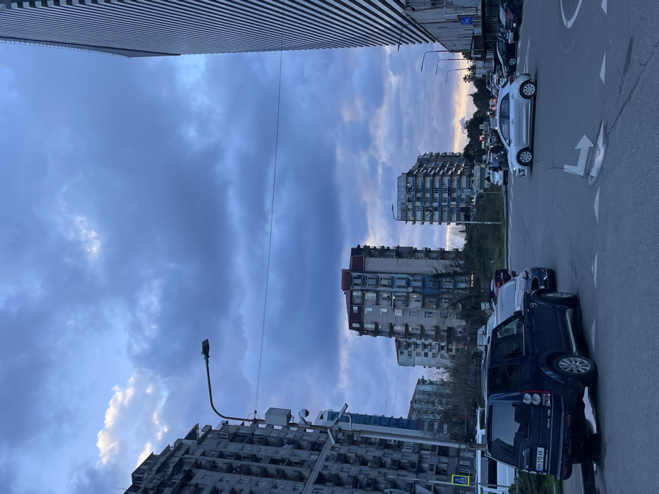 An outdoor wide shot depicts an urban streetscape in Batumi, Georgia, under an overcast sky with dark blue and grey clouds, suggesting dusk or dawn. In the foreground, a dark blue Land Rover Discovery 4 SUV, bearing Georgian license plate `KH-215-KK`, is parked on the left shoulder of an asphalt road, partially on a raised curb. To its right, in the middle of the road, a silver compact hatchback (possibly a Kia Rio) is positioned near an intersection or roundabout, with white directional arrows painted on the asphalt.
To the far left, a multi-story residential building with numerous balconies extends vertically, partially cropped from view, appearing weathered. A prominent street light pole equipped with multiple security cameras stands in front of this building. In the mid-ground, several other multi-story residential buildings with balconies are visible; one features a red roof and another shows distinct blue sections on its facade.
To the far right, a tall, modern building with a vertically striped facade, likely glass and paneling, is partially cropped. Overhead utility lines traverse the frame horizontally. Other vehicles are parked along the right side of the road and visible in the distance. Sparse greenery, including trees and bushes, is present between the road and the buildings in the middle ground. A pedestrian crossing sign is visible on the left, behind the parked Land Rover.