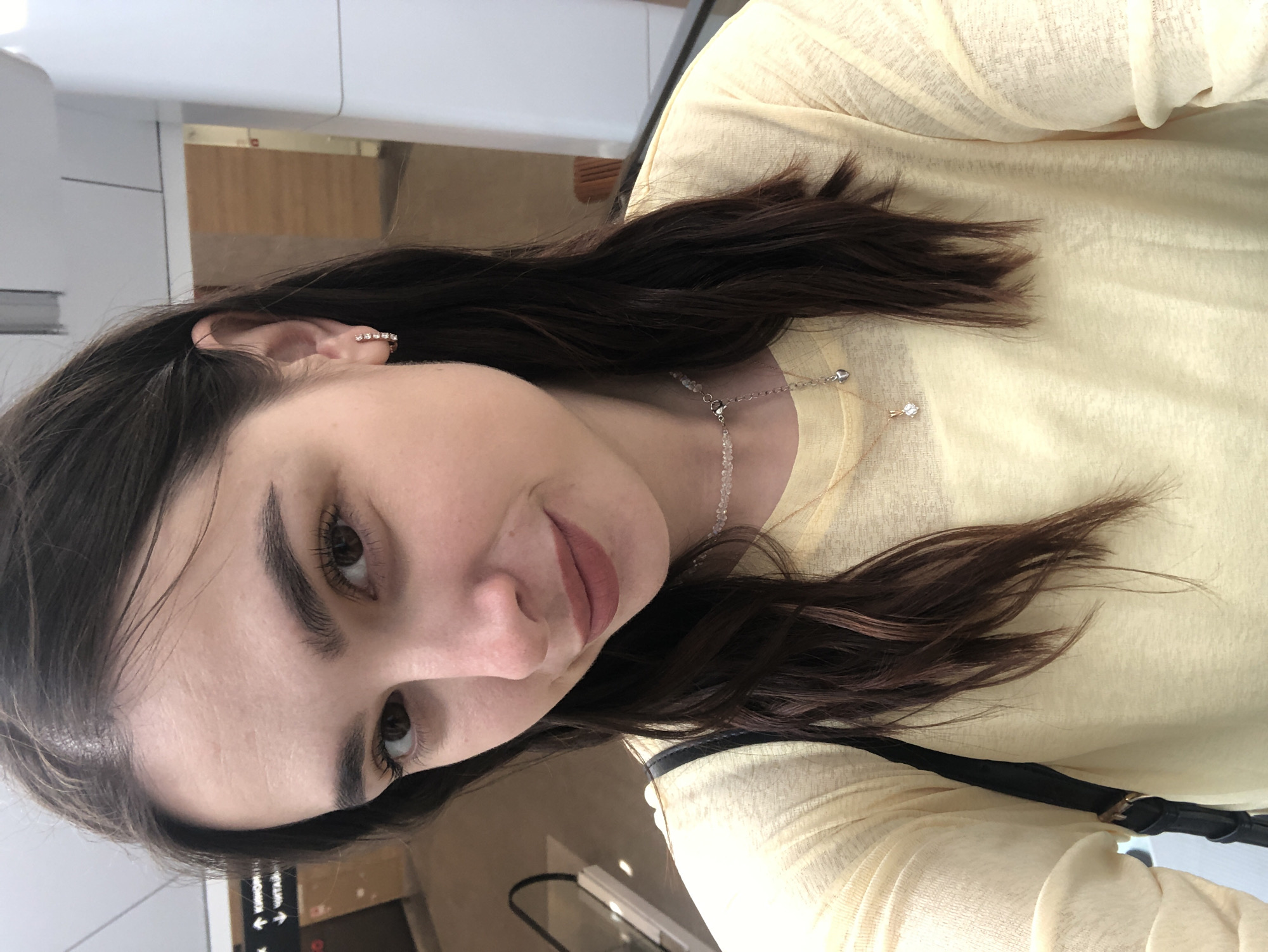 This is a medium, eye-level, portrait-style selfie of a young woman taken indoors. She has long, dark brown hair, fair skin, and brown eyes, with dark eyebrows. She is wearing a light yellow, long-sleeved, slightly sheer top. A delicate silver necklace with small clear beads and a small diamond pendant is visible around her neck. She is also wearing a small, sparkling earring on her right ear. Her lips are painted with a matte, muted rose lipstick, and she has a slight smile.
The background appears to be an indoor public space, possibly a shopping mall or an airport terminal. The lighting is bright and appears to be artificial. In the upper left of the image, a white structure is visible, and further back, wooden-paneled walls and some signage can be seen. A black strap, possibly from a bag, is visible across her chest. A hint of a black and white sign with some text is visible in the lower left portion of the image. The overall mood is relaxed and casual. The time of day and weather are not discernible from the image.