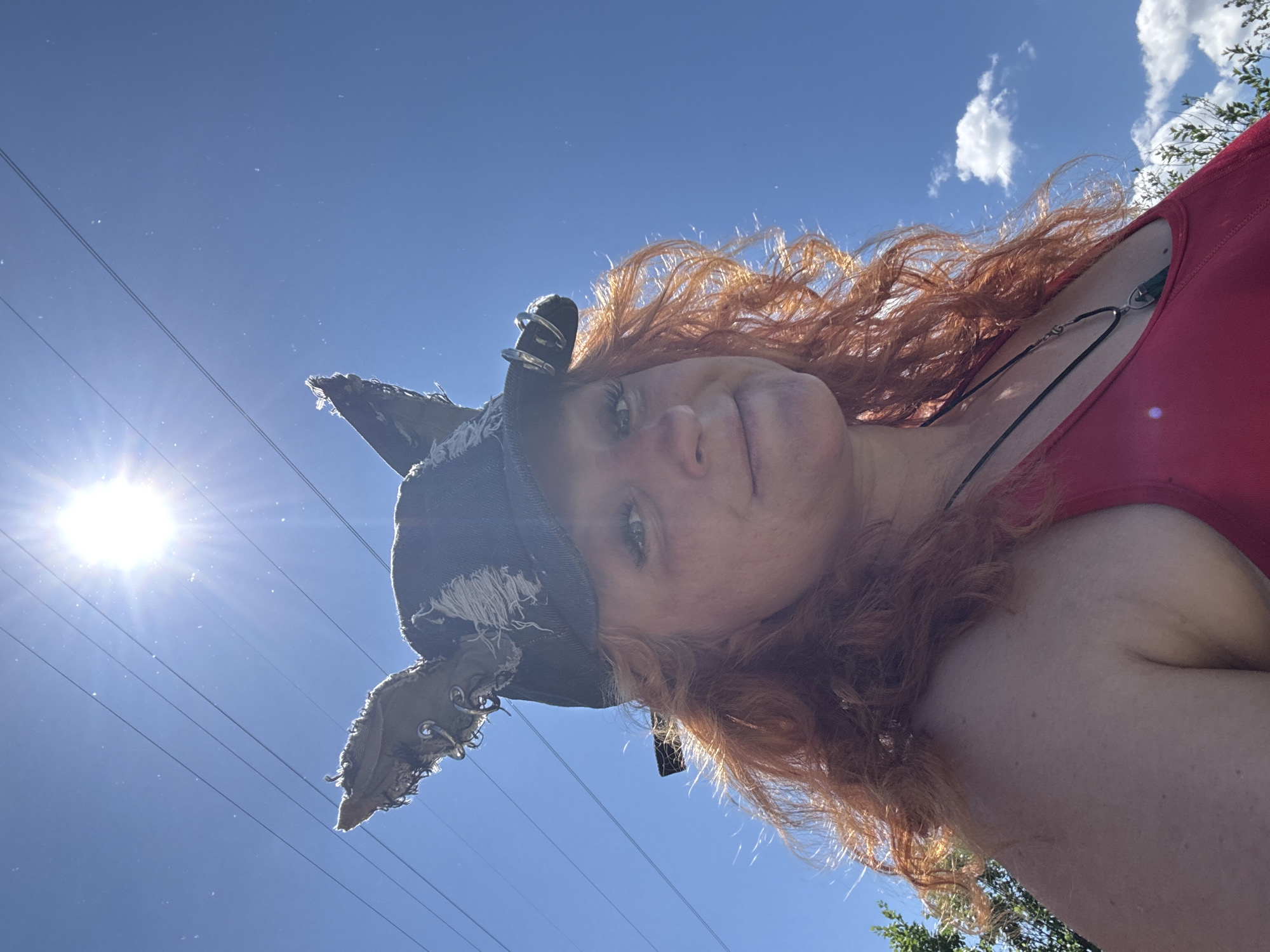 A woman with long, curly red hair and bright makeup poses outdoors against a bright blue sky with sun flares. She is wearing a red tank top and a black hat with distressed, pointed ear-like decorations made of fabric and metal rings. She is looking at the camera and smiling slightly. The background shows power lines and a hint of green foliage. The location is not specified as Chaikovsky, Russia, or any other city.