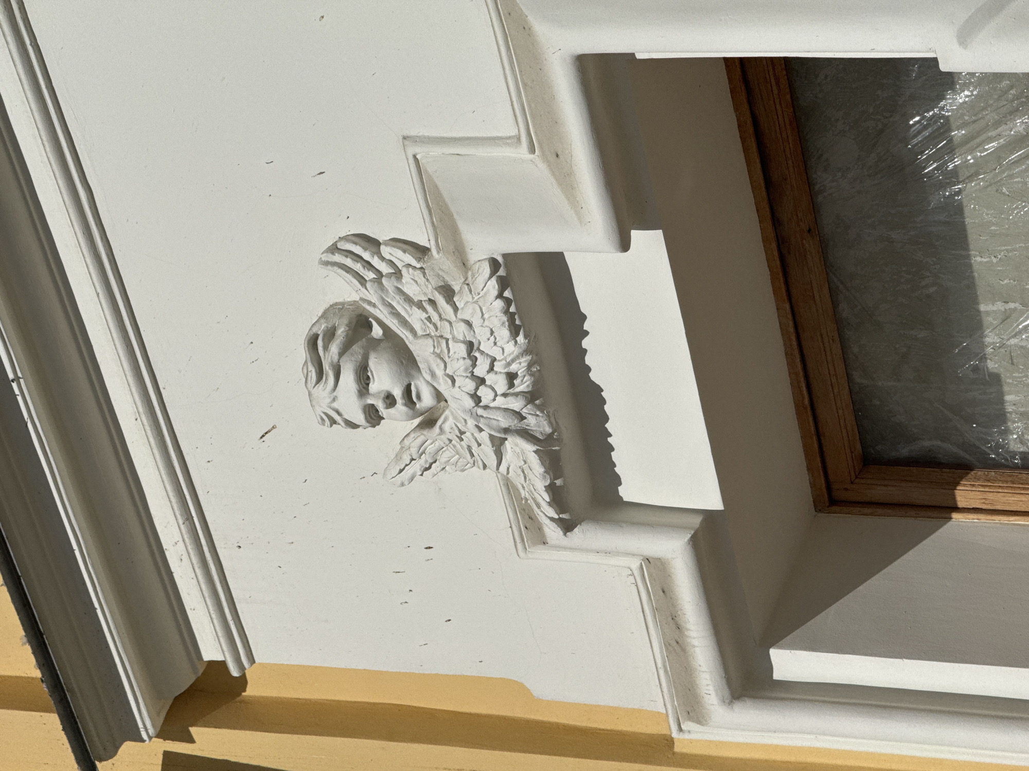 The media file captures a close-up, angled view of an architectural detail on a building in Saint Petersburg, Russia. The primary subject is a white, three-dimensional sculpture of a cherub or putto head with feathered wings, integrated into an elaborate white facade. The cherub's face is subtly detailed, looking upwards and to the right, framed by wavy hair and prominent wings that fan out behind its head.
The sculpture is set against a textured white wall with some minor specks and cracks, suggesting age. It is surrounded by sharp, geometric white molding that creates a series of stepped, L-shaped architectural elements. Strong sunlight from the upper right casts deep, angular shadows across these moldings and the wall, highlighting the contours and depth of the sculpture and the architectural features.
To the right of the cherub and molding, a portion of a window is visible, framed by warm brown wooden casing. The window pane itself is obscured by what appears to be clear plastic sheeting or film, possibly indicating renovation or protection. Below the white architectural elements, a strip of a yellow-painted surface is visible, contrasting with the white facade.
The image conveys a sunny daytime scene. There are no people or discernible activities, focusing solely on the static architectural beauty. No visible text is present.