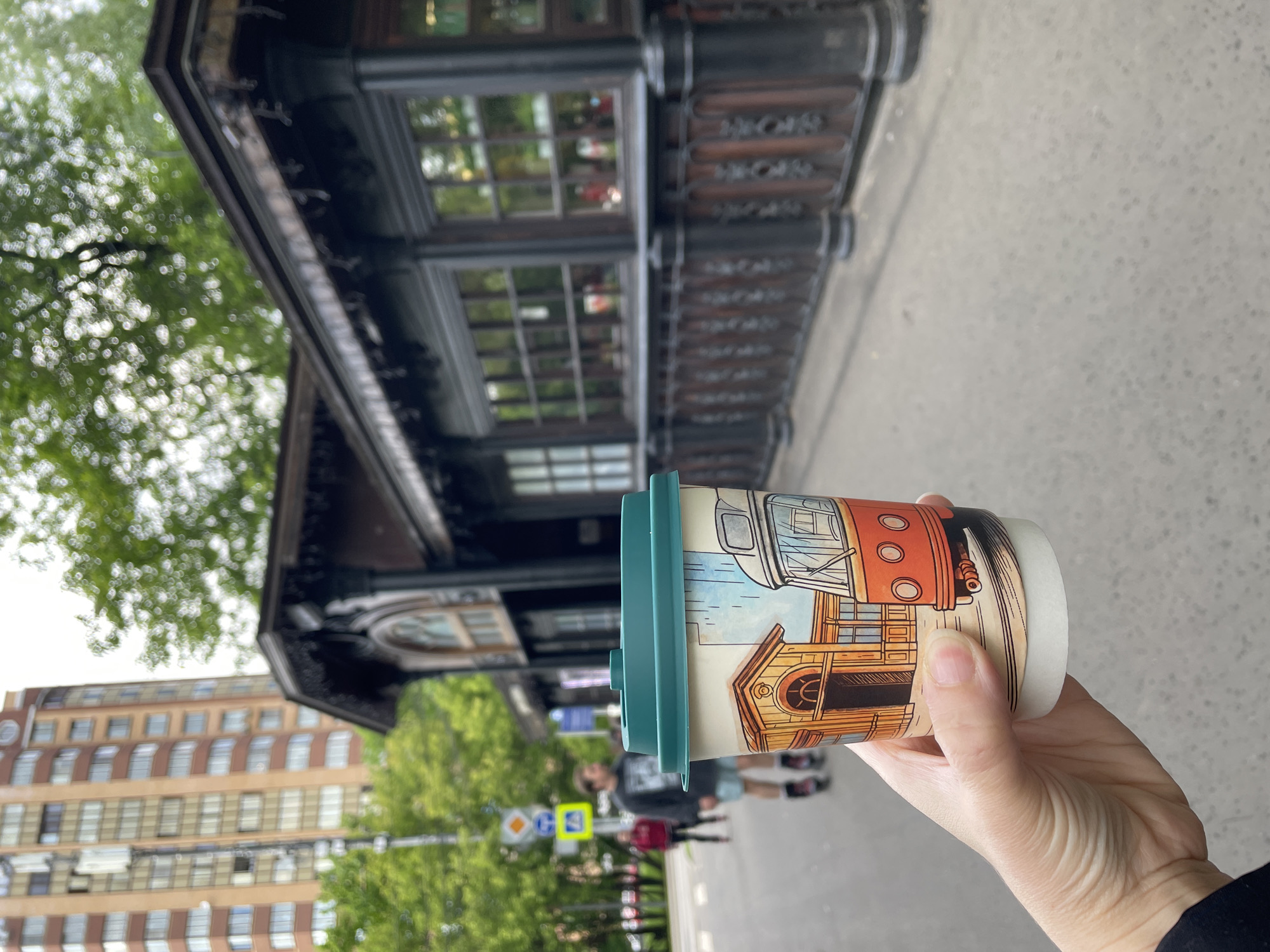 A person holds a coffee cup with a light green lid in their hand, with a scene of a city on the cup, in front of a building with a dark-colored, ornate facade, with multiple windows. Trees and a multi-story building with many windows are visible in the background. The photo was taken outside on a paved surface, likely a sidewalk or street. Pedestrians and traffic signs are further in the background. The cup has an illustration of a building and part of an orange-colored tram on it. The location is Khimki, Russia.