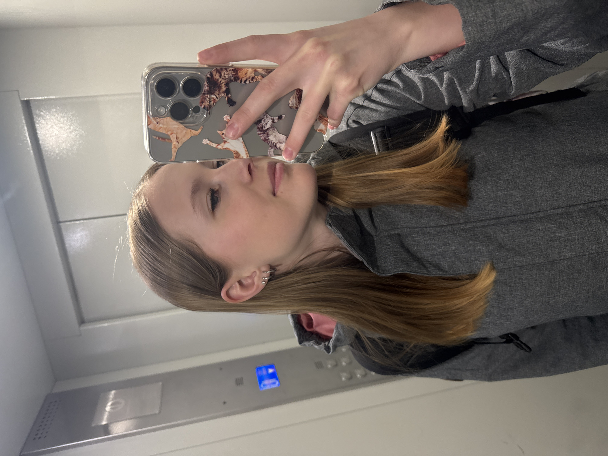 A fair-skinned person is taking a selfie in what appears to be an elevator. They are holding a phone with a cat-themed case in front of their face. The person is wearing a gray jacket, and a small part of a black backpack strap is visible. The elevator's control panel, with blue illuminated buttons, is visible in the background, along with the metal elevator walls. The location cannot be determined as Chaikovsky, Russia, based on visual information alone.