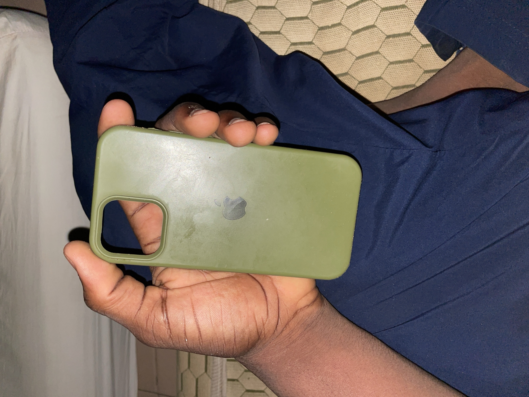 A person's hand, with dark skin and visible fingerprints, is holding a green iPhone case. The case has a cut-out for the camera lens. The background shows dark blue fabric and a light-colored textured surface. The image appears to be taken indoors, possibly in a bedroom, and no specific location within Safari, Nigeria, is discernible.