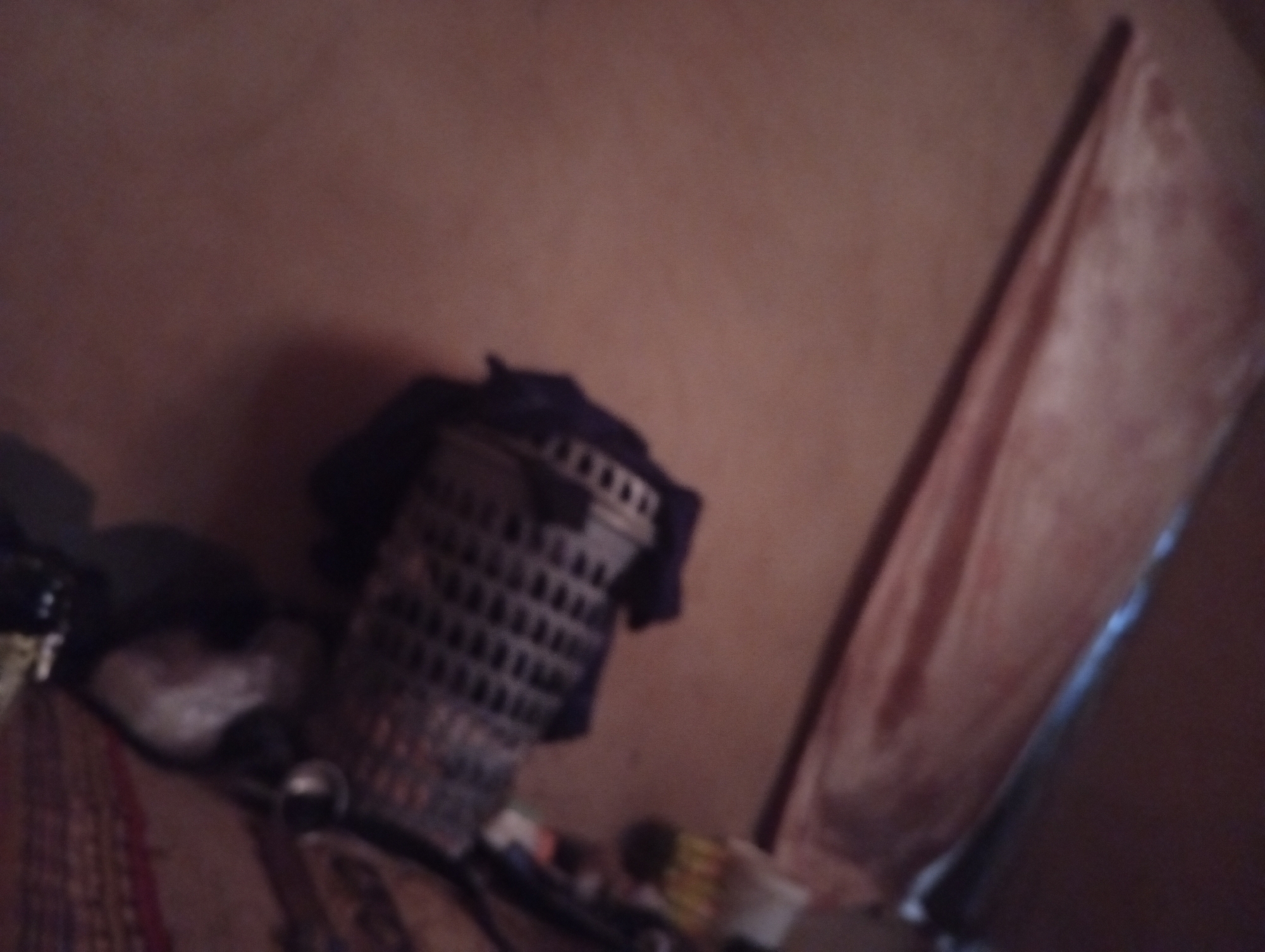 The image shows a cluttered interior, likely a room. A perforated plastic basket sits on the floor, partially covered by a dark fabric. Scattered around the basket are various objects, including what appears to be a white plastic container and other unidentifiable items. To the right of the basket is a door leaning against the wall. There are no people present in the image.
The image is taken from Rigasa, Nigeria.