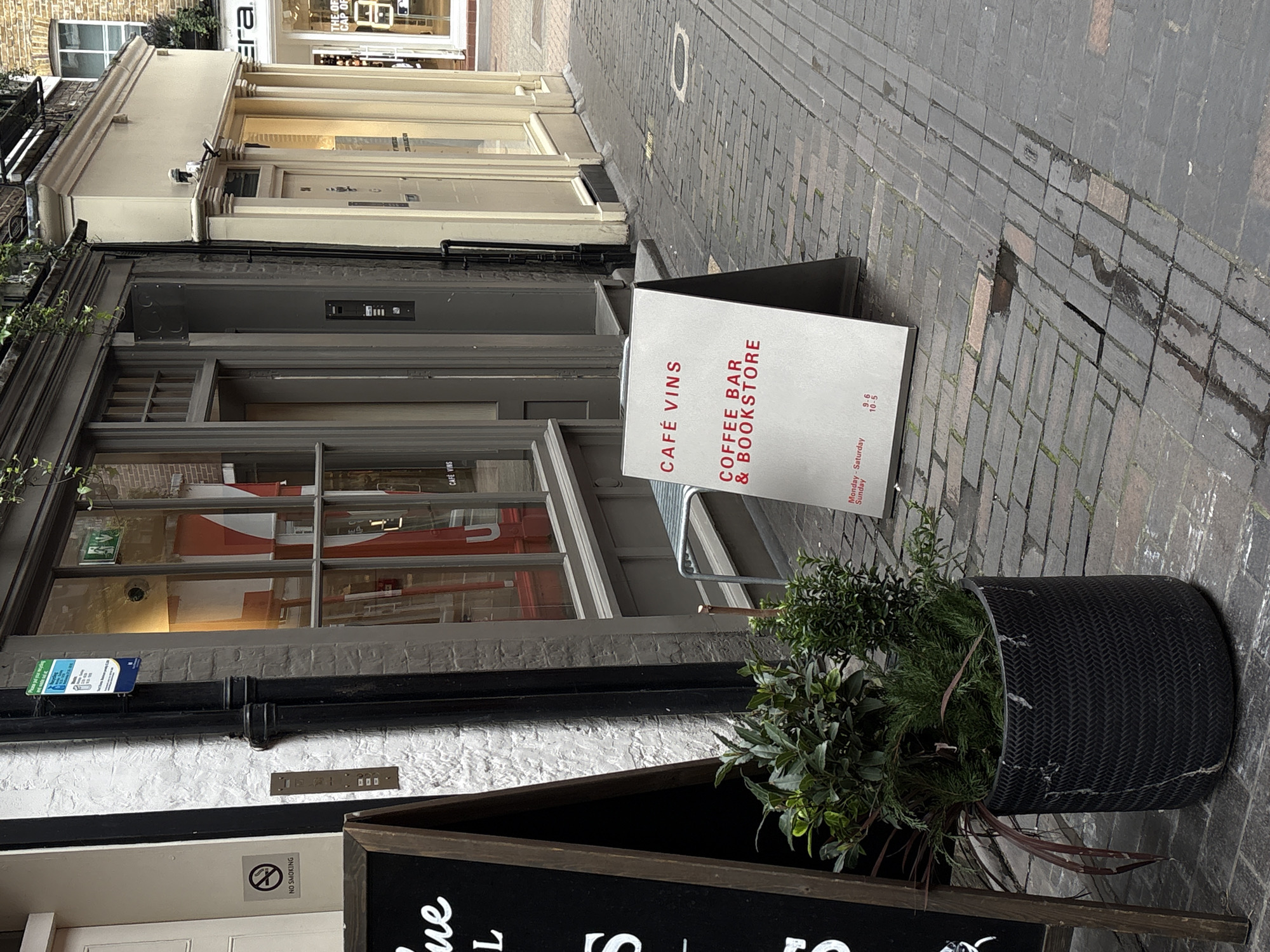 Here is a detailed description of the image:
The photograph depicts a street scene in London, United Kingdom. The camera is tilted slightly to the right. The street is paved with gray bricks.
On the left side of the frame is the façade of a building. There is a sign that reads "CAFE VINS COFFEE BAR & BOOKSTORE." A black pipe runs horizontally across the white lower portion of the building. There is a "NO SMOKING" sign.
In the foreground, there's a large black woven planter filled with greenery. Behind the planter is another sign advertising the Cafe Vins and its menu of “ue L S”.
The building next door on the left side of the frame has a store front with the logo "Era THE OF CAP OF".
The street appears relatively empty, with no visible people.