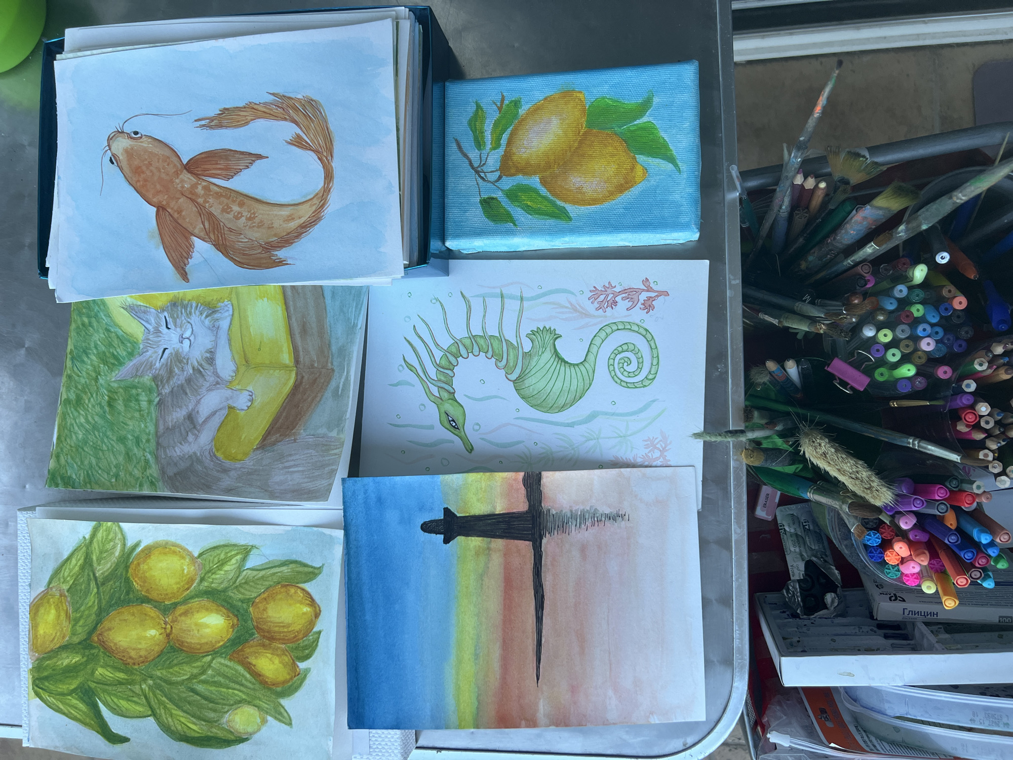 This overhead image displays a collection of art pieces and art supplies arranged on a metallic surface, likely a table or desk, in Baošići, Montenegro.
The main subjects are six distinct artworks. In the top left, a watercolor painting depicts a golden-brown fish, possibly a koi or goldfish, swimming in light blue water. Above the center, a small canvas features two yellow lemons with green leaves on a light blue background. Below the fish, another watercolor shows a gray tabby cat peacefully sleeping on a yellow object, with green foliage in the background. Centrally located, a stylized green sea creature, resembling a spiky seahorse or sea dragon, is rendered in fine lines and washes against a pale blue background with abstract red and green underwater flora. In the bottom left, a watercolor presents a cluster of bright yellow lemons and lush green leaves. The final painting, in the bottom center, features a dark, abstract cross-like structure centered over a horizontal watercolor gradient transitioning from blue to yellow, orange, and pale pink, with a reflection indicated below the structure.
To the right of the artworks, various art supplies are visible, including a container overflowing with numerous colored markers, brushes, and colored pencils. Other items include an eraser and a white paint palette with traces of paint.
Notable details include the variety of art mediums, predominantly watercolor for the paper pieces and possibly acrylic for the small canvas. The brush strokes and detailing vary across the pieces, showcasing different artistic styles. One box of art supplies prominently displays the Cyrillic text "ГЛИЦИН" (GLITSIN) and "АКВАРЕЛЬ" (AKVAREL), indicating watercolors. A white sticker shows a partial date "2022.13.46". The overall scene suggests an artist's workspace or a display of completed works.