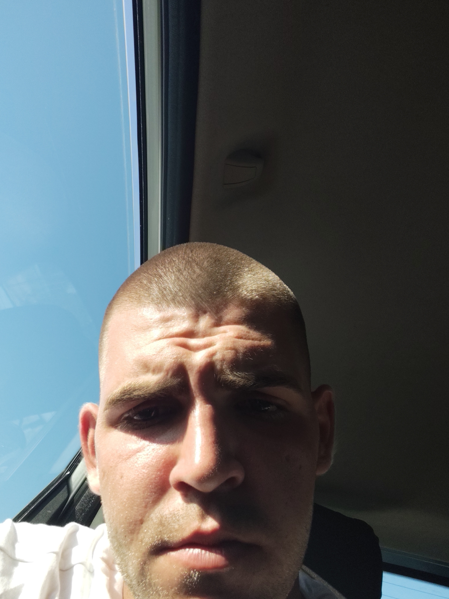 A close-up, eye-level selfie depicts a fair-skinned man with a buzz cut and a short beard. He is positioned centrally, with his face occupying most of the frame. The man's expression is neutral, and his eyebrows are furrowed, suggesting concentration or concern. He is wearing a white t-shirt. The background is divided into two distinct areas: on the left, a bright blue sky is visible through a window, and on the right, the dark interior of a vehicle, possibly a car, is apparent. A sun glare illuminates the left side of the man's face. The image was taken outdoors, likely inside a vehicle in Carrù, Italy.