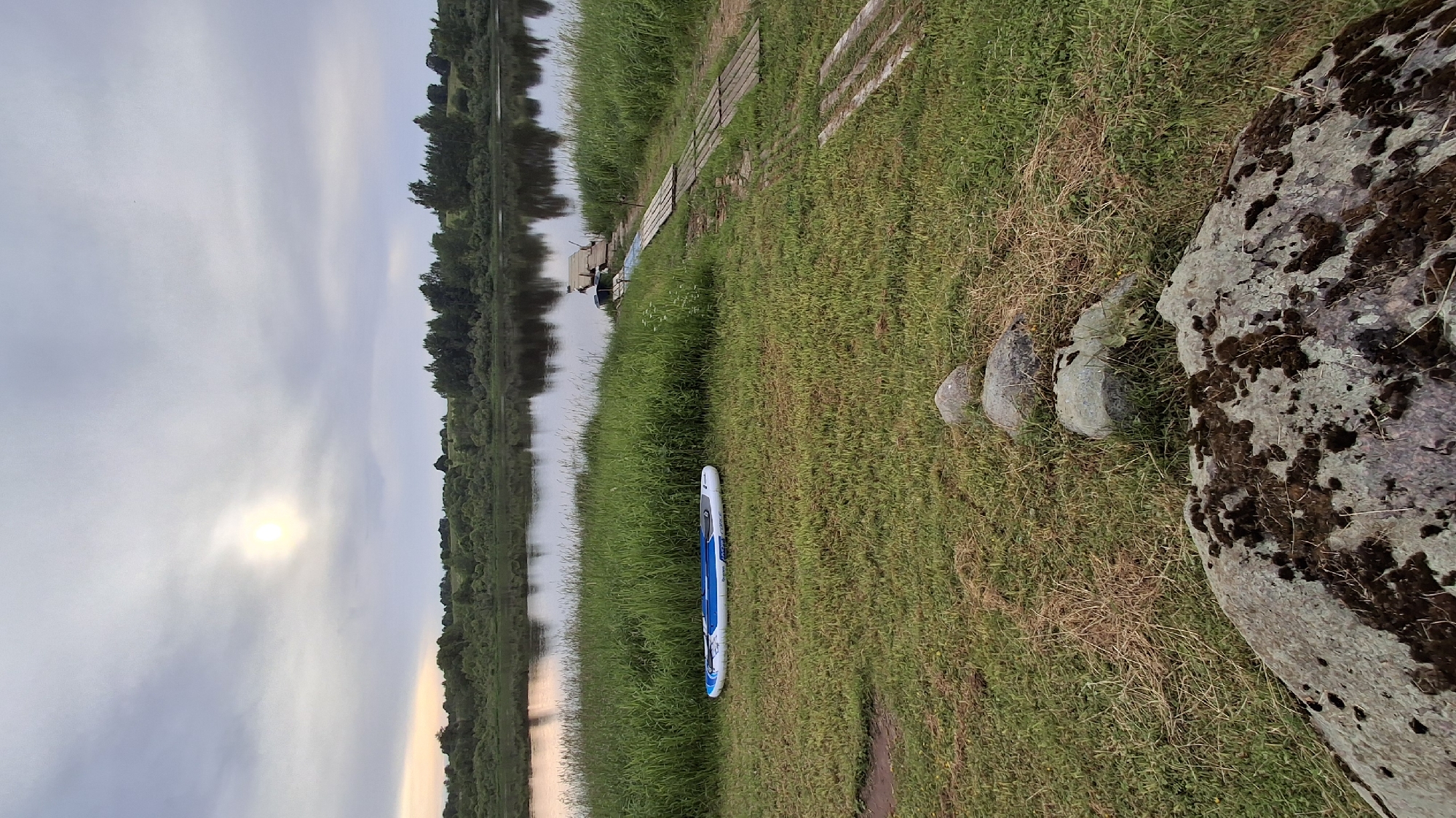 This media file depicts a serene waterside scene in Sitno, Russia. The primary subject is a blue and white stand-up paddleboard lying on a grassy bank beside a calm body of water. The water reflects the overcast sky and the lush green trees lining the opposite bank, suggesting a natural, undeveloped setting.
In the background, a wooden walkway or jetty extends into the water, with a few small boats moored nearby. Three smooth, rounded stones are arranged on the grass near a large, moss-covered boulder, adding a touch of natural artistry to the scene.
The time of day appears to be late afternoon or early evening, given the soft, diffused sunlight peeking through the clouds and the warm hues near the horizon. The weather is cloudy, contributing to the tranquil atmosphere. The image evokes a sense of peace and quietude, perfect for outdoor recreation or contemplation. There is no visible text in the media file.