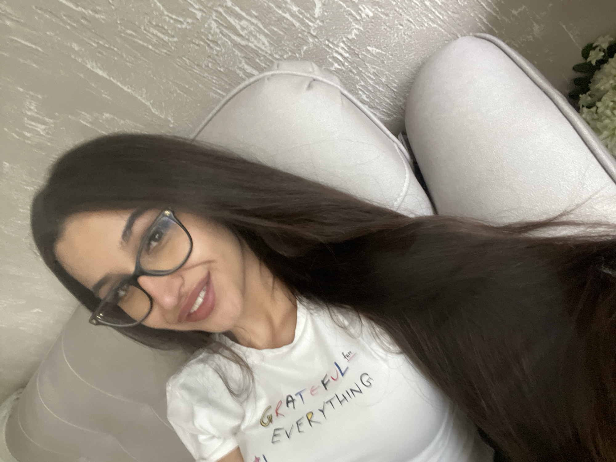 A young woman with long, dark hair and glasses smiles while lying on a light-colored couch. She wears a white t-shirt with the words "GRATEFUL EVERYTHING" printed on it. Part of a white flower arrangement is visible in the background. The location appears to be indoors, with a textured, neutral-colored wall. The city and state of Unknown, Unknown, cannot be confirmed from the image.