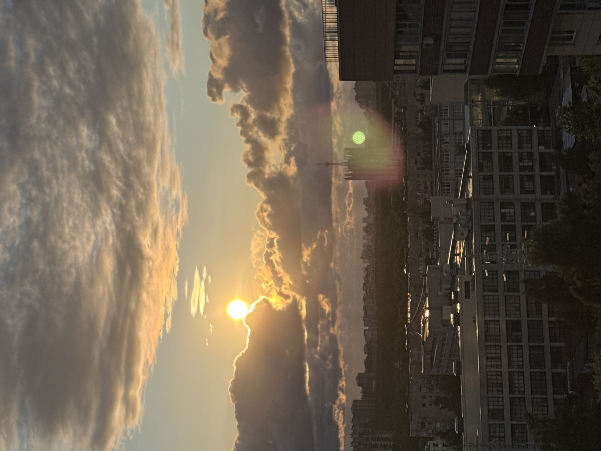 An aerial view captures a cityscape of Sinyavino, Russia, at sunset. The sky is filled with dramatic clouds, illuminated by the setting sun which peeks through the cloud cover, casting a warm, golden glow. In the foreground, several multi-story buildings with visible windows and balconies dominate the scene. A prominent building on the right features a dark brick facade and a metal railing. In the middle ground, a silhouette of a tower-like structure is visible, with a green circular light reflection in front of it, possibly a lens flare or atmospheric phenomenon. Beyond the buildings, a dense forest stretches across the landscape, leading to a distant urban skyline. The overall impression is one of a city transitioning from day to night, with the natural beauty of the sunset contrasting with the man-made environment.