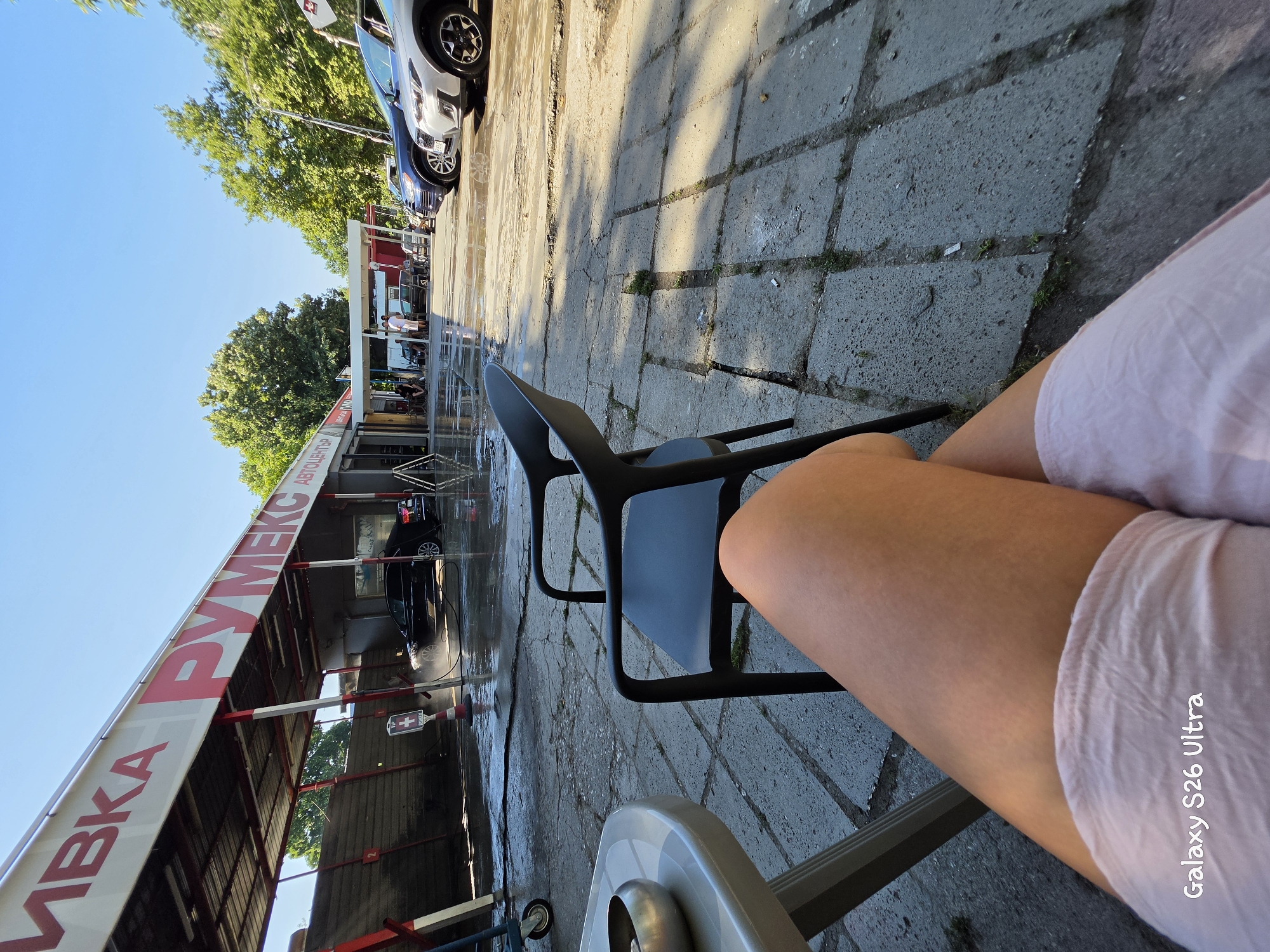 A low-angle shot captures a sunny outdoor car wash in Plovdiv, Bulgaria, with a person's legs in the foreground. The car wash features a red and white sign with Cyrillic lettering reading "АВТОЦЕНТЪР РУМЕКС" (Autocenter Rumex). Several cars are visible, some being washed, others parked. The ground is wet and reflective, indicating recent activity. A black plastic chair sits unoccupied in the middle ground, to the left of the frame. Lush green trees line the background, suggesting a warm, pleasant day. The image includes the watermark "Galaxy S26 Ultra" in the bottom left corner.