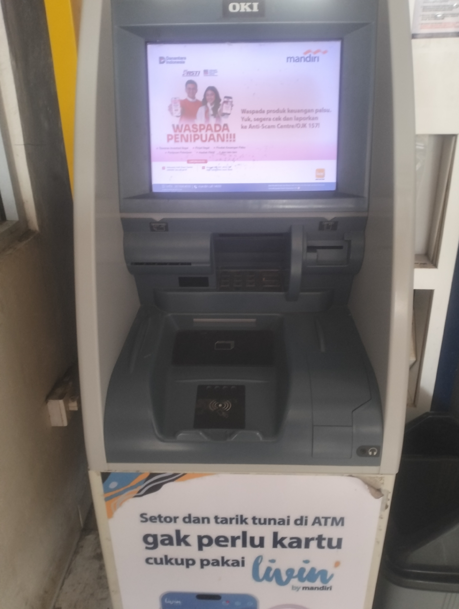 A close-up, front-facing shot shows an OKI ATM machine. The screen displays a pink and white advertisement for Mandiri Bank, featuring a couple holding smartphones and the text "WASPADA PENIPUAN!!!" (Beware of Fraud!!!). Below the screen, the ATM has a card slot, a keypad, a cash dispenser, and a contactless payment reader. A white poster is attached to the bottom of the ATM, with text in Indonesian that translates to "Deposit and withdraw at the ATM, no card needed, just use Livin' by Mandiri." The ATM is situated indoors, against a textured wall with yellow and white vertical stripes. The floor is not visible. The overall scene appears to be a public area, likely a bank branch or a commercial establishment in Dumai City, Indonesia, given the Indonesian language on the advertisement and poster.