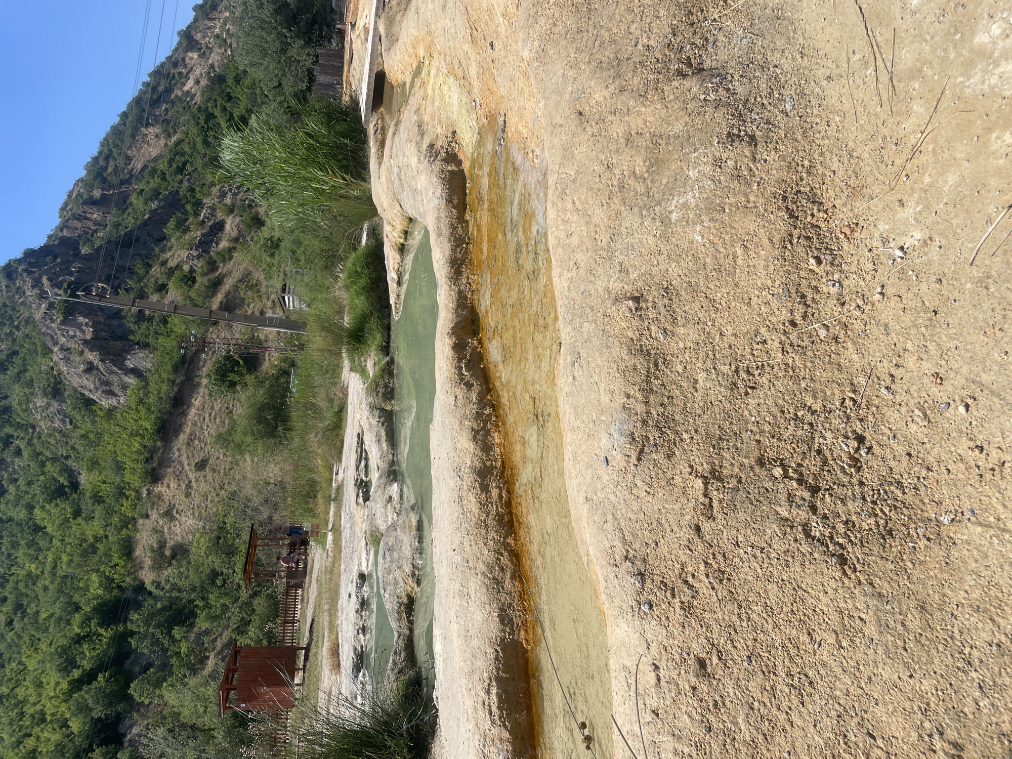 This image depicts a natural outdoor thermal springs area located in General Todorov, Bulgaria, under a clear, bright blue sky. The foreground features dry, light brown, gravelly ground with scattered small rocks and sparse dried plant debris. Two distinct, shallow, irregular pools of yellowish-brown water with orange mineral staining along their edges are prominent.
Moving into the midground, multiple larger, irregularly shaped pools containing milky light green water are visible, embedded within naturally formed light brown and beige rock terraces. Clumps of tall, vibrant green reeds or grasses grow on the left and right sides of these pools. To the far right, a narrow wooden plank bridge crosses a water channel.
In the midground-left, behind the green pools, a small, open-sided wooden shelter or gazebo provides shade, under which two individuals are present: one person is standing, wearing a blue top and dark bottoms, while another person is seated, wearing a light-colored top and dark bottoms, seemingly resting. Adjacent to the shelter on the left is a reddish-brown wooden structure, resembling an outdoor changing facility or toilet. A wooden fence runs behind these structures, delineating a dirt path.
The background is dominated by a steep hillside or mountain heavily covered with dense green trees and vegetation. Portions of exposed rock faces are visible on the upper right side of the mountain. Utility poles with overhead power lines are positioned against the hillside on the right. The scene is brightly lit by direct sunlight, indicating a clear, sunny day.