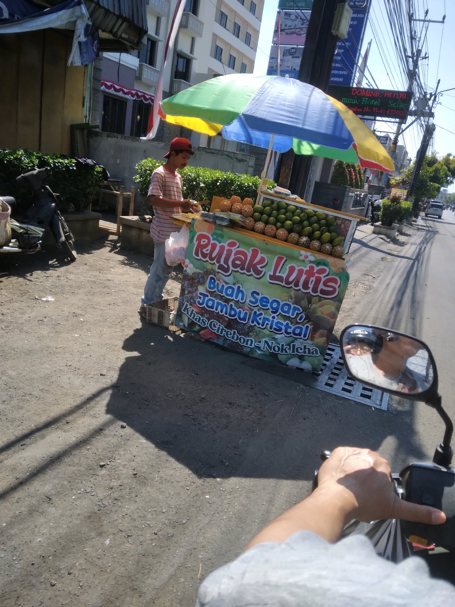 An Indonesian man in a red cap and striped shirt stands behind a fruit cart, preparing a sale. The cart is laden with various fruits, including mangoes and what appear to be pineapple-like fruits, and is adorned with a colorful banner that reads "Rujak Lutis Buah Segar, Jambu Kristal, Khas Cirebon-Nok Icha". An open umbrella shades the cart. In the background, buildings and telephone poles are visible, suggesting an urban setting. A motorcycle is parked to the left of the fruit cart, and a motorcycle's side mirror, reflecting the rider and the scene, is in the foreground. The image appears to be taken in Purwokerto, Indonesia, based on the sign referencing "Cirebon".