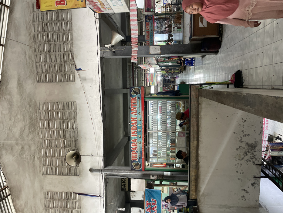 This image captures an indoor market or shopping area within Nganguk, Indonesia. The scene is characterized by a concrete structure featuring a high ceiling with exposed beams, multiple rectangular ventilation blocks, and a large, convex security mirror mounted high on a pillar. The flooring consists of light-colored tiles.

In the immediate foreground, a concrete barrier with a sloping section indicates a lower level, where hanging textiles and a metal staircase are partially visible. To the left, a woman wearing a grey long-sleeved top and a dark headscarf stands near a stall, partially obscured by a blue sign.

Central to the midground is a prominent stall with a green frame and a dark blue sign displaying "JEPARA INDAH ANTIK," an address "Banyuputih Rt. 00, Rw 02 Kec. Kalinyamatan Jepara," and a phone number. This stall exhibits numerous identical silver cylindrical bottles on multiple shelves, with a lower row of dark-colored bottles. Two males are positioned behind the counter; one in a red cap is looking down at a device, while the other is partially obscured.

To the right, a tiled aisle extends into the background, flanked by various other stalls. A broom and dustpan rest on the floor near a structural pillar. Further along the right, another stall, also identified as "JEPARA INDAH ANTIK" by a partially visible sign, displays an array of smaller items. A white megaphone is mounted on a pillar in the midground. A young girl in a pink headscarf and a long pink dress walks from the background towards the foreground-right, looking to her right. In the deeper background, several indistinct figures, including one seated on a blue stool, are visible among other market stalls.