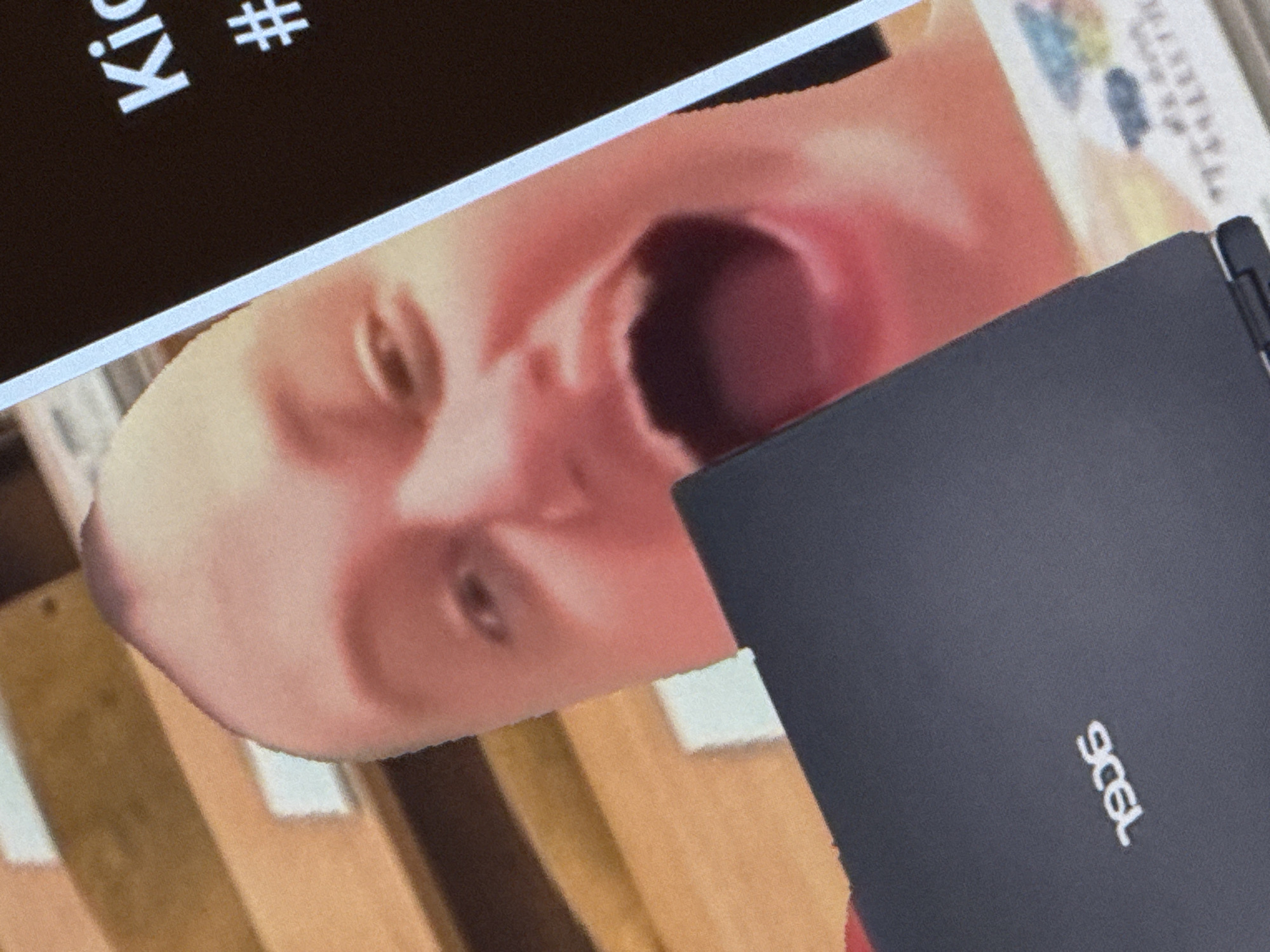 The image shows a close-up of a person's face, which is largely out of focus, but their mouth is wide open in a scream or shout. The person is holding a dark-colored laptop in front of them. The laptop has a logo that appears to be "1906". In the background, there are wooden shelves with some white objects on them. To the right of the person's face, a dark screen with white text is visible. The text reads "Ki" and "#". The scene appears to be indoors, possibly in a room or a store. There is no identifiable information about the location in Saratov, Russia.