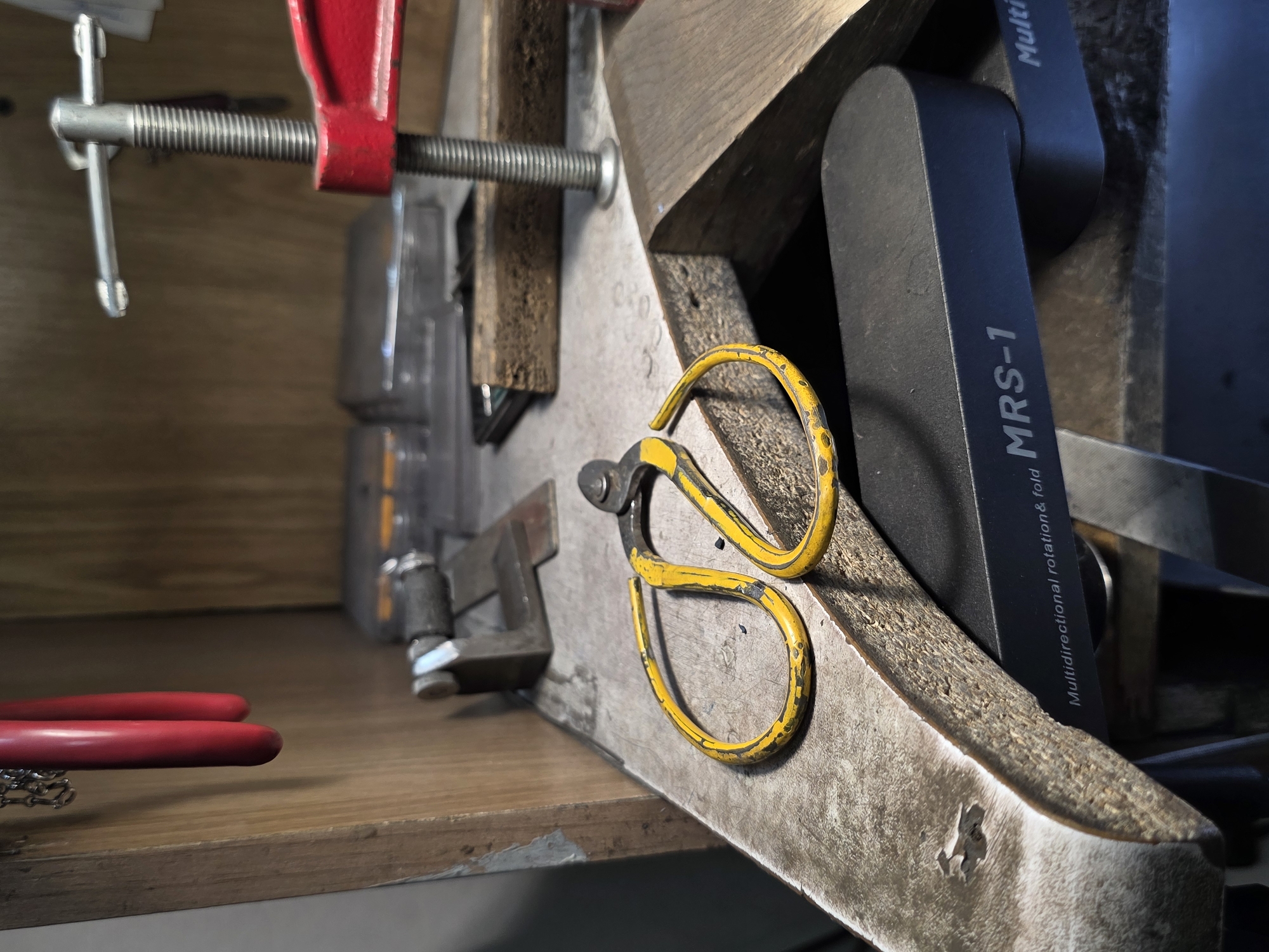 The image depicts a close-up view of a workbench, likely in a workshop or studio. The workbench is cluttered with various tools and materials, suggesting a workspace for crafts or repairs. In the foreground, a pair of yellow-handled wire cutters with a black pivot is prominently displayed. The cutters are open and resting on a textured metal surface, possibly part of a larger workbench or a bench vise. To the right, a dark, curved object with the inscription "MRS-1" and "Multidirectional rotation & fold" is visible, suggesting it's a rotating base or holder.
Behind the wire cutters, a variety of objects are present. A red clamp is attached to a threaded rod, likely holding something in place. Other items include what appear to be small metal blocks, and some transparent containers with contents that are not clearly discernible. The background shows wooden panels, possibly shelves or the back of a cabinet, with a chain and red handles hanging from the top left. The overall impression is one of a functional, actively used workspace.
Regarding the location in the city of Kazan, Russia, there is no visual information within the image that directly or indirectly indicates its presence in Kazan. The scene is generic to many workshops or craft areas.