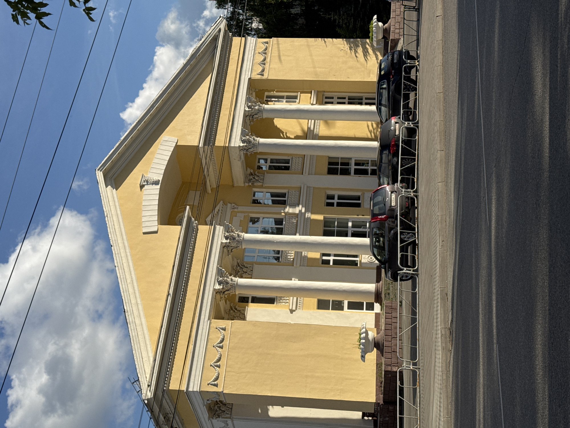The image shows a building with a yellow facade and classical architectural elements, including large white columns and decorative moldings. The building appears to be a public institution, possibly a theater or a museum, given its grand design. The facade features multiple stories with rectangular windows, some of which reflect the blue sky and white clouds. At the top, a triangular pediment suggests a neoclassical style.
In front of the building, several black cars are parked along the sidewalk, separated from the road by a white metal fence. The cars are parked parallel to the building, and some have license plates visible, though the text is not legible. A sidewalk runs along the front of the building, and beyond that is an asphalt road with a white line marking a lane. The ground is bathed in sunlight, casting long shadows from the fence and the cars onto the road, indicating it is likely daytime.
The scene is set against a bright blue sky with fluffy white clouds, suggesting fair weather. Overhead, power lines stretch across the frame, adding a touch of modern infrastructure to the otherwise traditional architectural setting. In the upper left corner, the leaves of a tree are visible, casting dappled shadows on the power lines.
There are no people visible in the image, nor any explicit activity taking place other than the presence of parked cars. The overall impression is one of a calm, sunny day in a city, with a focus on the striking architecture of the building. There are no discernible emotions conveyed, and the location cues are subtle, with the mention of Izhevsk, Russia, providing the geographical context.