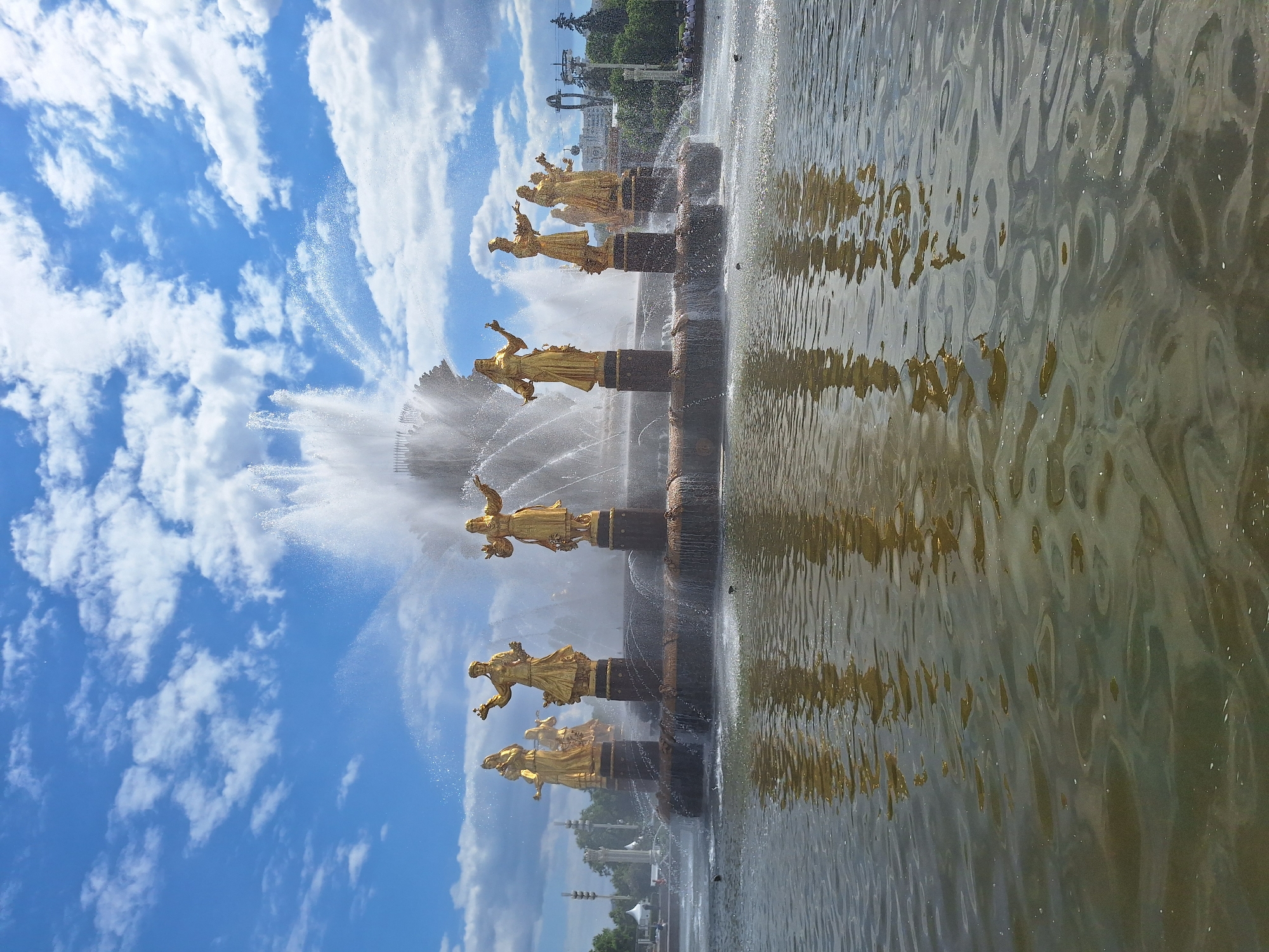 The image captures the iconic "Friendship of Nations" fountain located at VDNKh (Exhibition of Achievements of National Economy) in Moscow, Russia, under a bright, sunny sky.
The central subjects are a series of golden statues arranged in a line within a large fountain basin. There are at least eight distinct golden figures visible, each standing on an individual dark-colored pedestal. These statues depict female figures, likely representing the former Soviet republics, dressed in traditional attire and appearing to hold symbolic items such as sheaves of wheat. Their polished golden surfaces reflect the sunlight, standing out against the surrounding water and sky.
Powerful streams of water erupt upwards and cascade around the statues, creating a dynamic display. The water sprays high into the air, forming a misty halo and adding to the grandeur of the fountain. The fountain's large pool occupies the lower portion of the image, its surface rippled by the water's movement. This creates shimmering reflections of the golden statues and the blue sky, with the patterns of light and dark on the water's surface.
The setting appears to be a clear and pleasant day, indicated by the vibrant blue sky dotted with scattered white, cumulus and cirrus clouds. In the background, beyond the fountain, hints of green trees and distant architectural structures can be observed, suggesting a spacious park or exhibition ground. The overall impression is one of historical grandeur and artistic craftsmanship. There is no visible text in the image.
