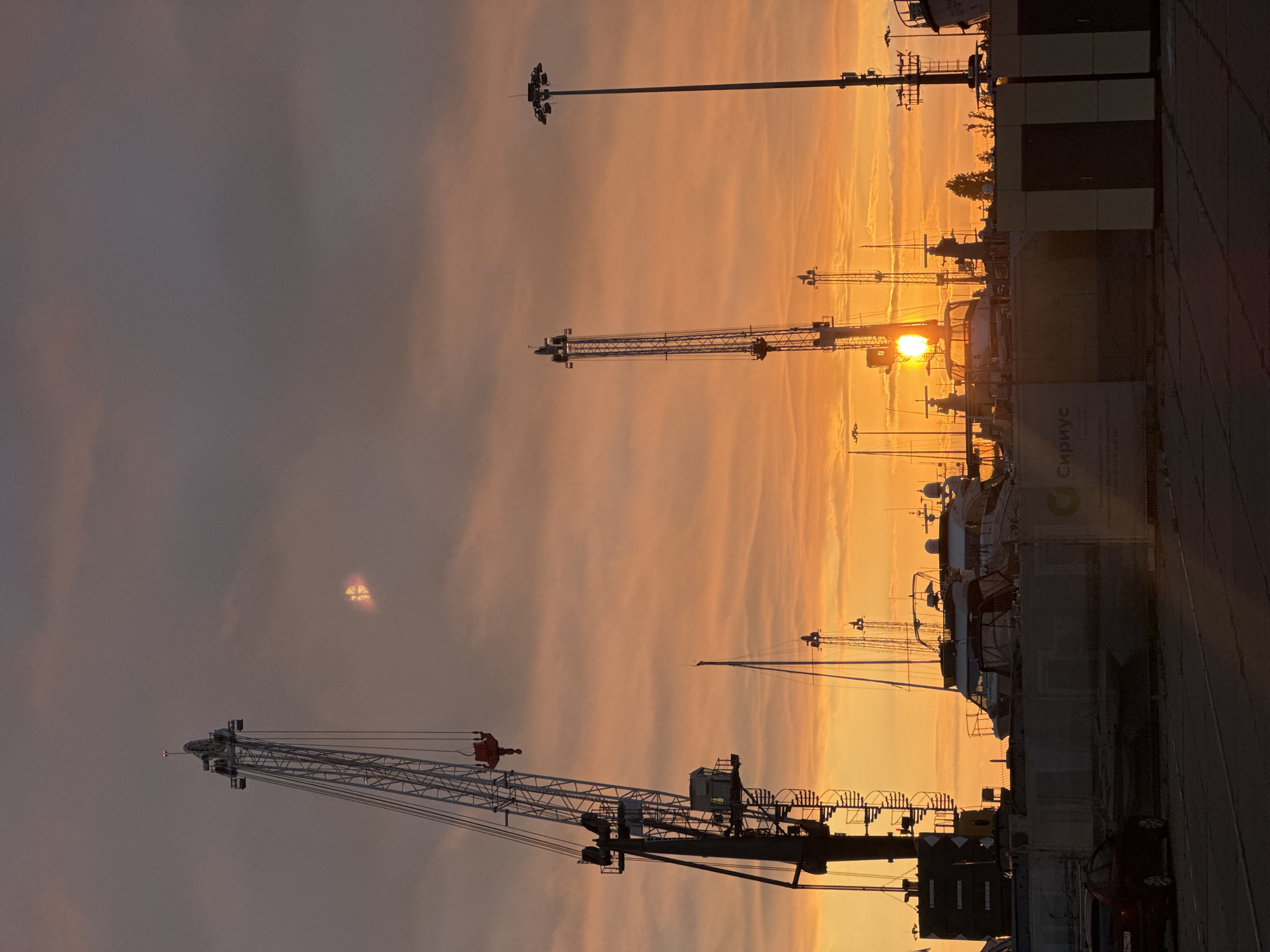 The image depicts a harbor scene in Sirius, Russia, during sunset. The sky is a vibrant gradient of orange and pink, with scattered clouds. Silhouetted against the sky are several large industrial cranes, one of which is prominently featured in the foreground on the left. The cranes are metallic gray and white, with visible gantries and cables.
In the middle ground, several docked yachts and boats are visible, their masts and structures also silhouetted. Some of these appear to be luxury vessels. In the background, there are more industrial structures, including tall light poles.
The foreground shows a paved area, possibly a dock or road, with a dark-colored car parked on the left. A low wall or barrier runs across the middle ground, with some advertising or signage visible on it, including the word "Сириус" (Sirius) in Russian. The overall impression is one of a working port or marina at the close of day, with the dramatic colors of the sunset creating a striking visual contrast with the industrial elements. There are no people clearly visible in the image.