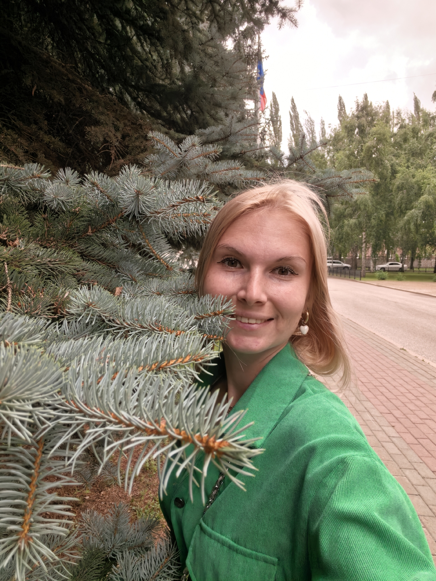 The media file is a selfie of a young woman with blonde hair and light skin, taken outdoors in Sterlitamak, Russia. She is smiling and wearing a vibrant green jacket. Her right side is partially obscured by the branches of a blue spruce tree, which dominates the left side of the frame. Her earrings are visible, one of which is shaped like a white heart.
Behind her, a paved walkway is visible, lined with trees. Beyond the walkway, a street with parked cars and more trees can be seen. In the distance, a flag with red, white, and blue colors is flying, possibly a Russian flag. The sky is overcast, suggesting a cloudy day. The overall atmosphere is casual and relaxed, with the woman appearing happy and enjoying the outdoors. The image is likely taken during daylight hours.