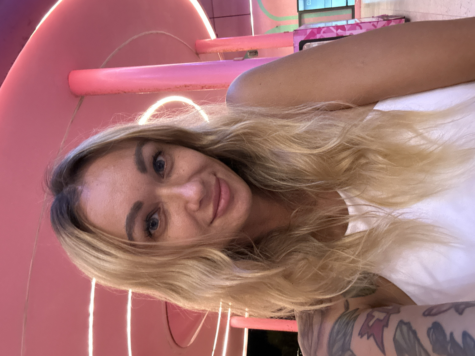 The image is a close-up portrait of a young woman with long, wavy blonde hair and bright blue eyes, smiling faintly at the camera. Her face shows freckles, and she wears a white top. A tattooed arm is visible in the lower right portion of the frame, featuring intricate designs including what appear to be floral, leaf, and abstract patterns in dark and reddish tones.
The background depicts a modern and vibrant interior, dominated by various shades of pink. Curved pink structures, resembling decorative tubes or archways, are brightly illuminated by white strip lighting that creates a glowing effect. Further in the background, additional structures combine pink and light green elements. The artificial lighting suggests an indoor setting or an evening shot. The woman appears happy and relaxed. There is no visible text in the image. The location context is Tien Thanh Ward, Vietnam.