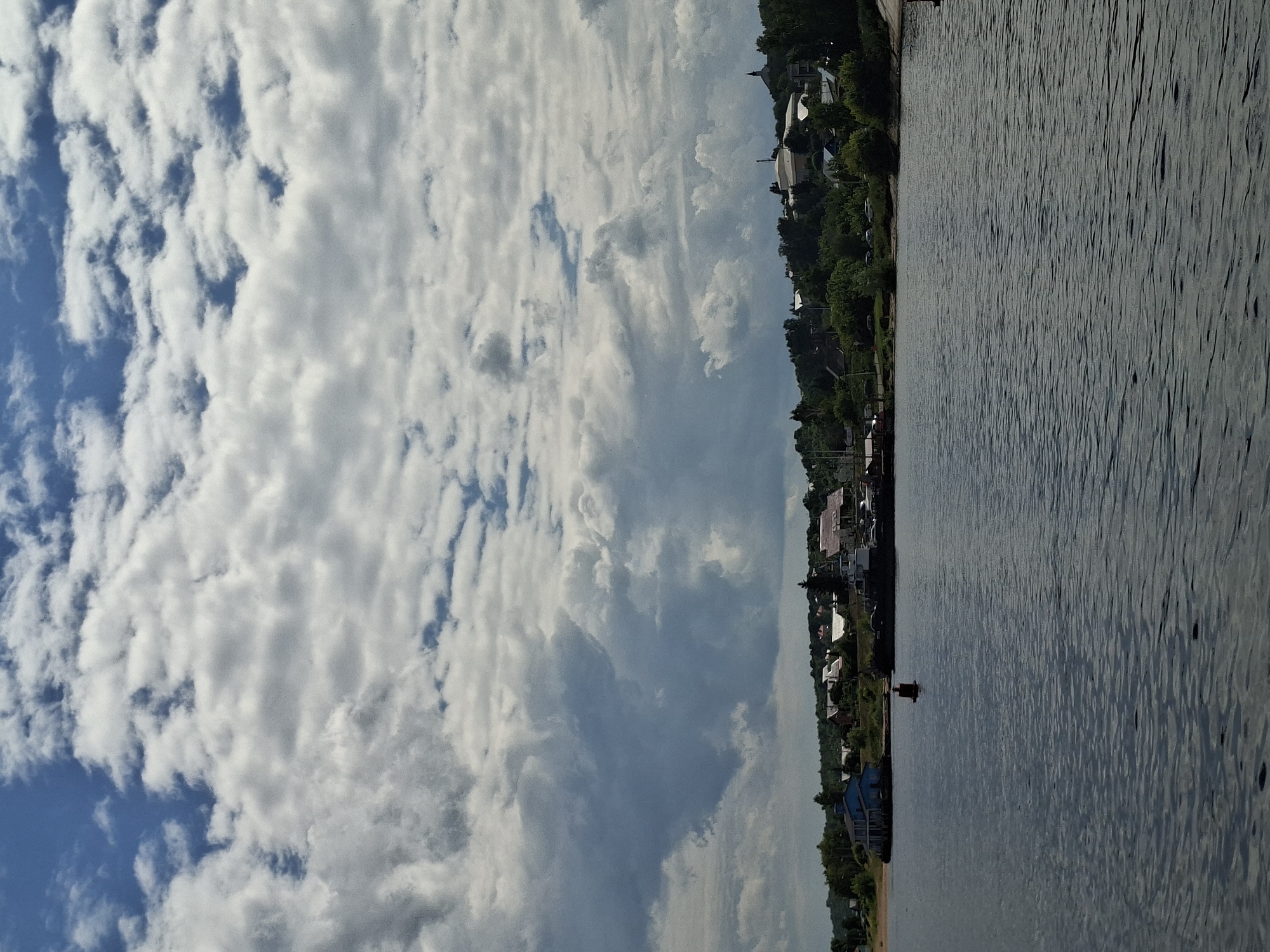 The image depicts a wide, dark body of water in the foreground, with gentle ripples on its surface. Along the far bank, a low-lying settlement is visible, characterized by a cluster of buildings with red and white roofs, interspersed with lush green trees. A prominent blue building stands out on the left side of the bank, near a sandy patch. Further to the right, several boats are docked along the waterfront. Above the settlement, a hilly landscape stretches into the distance, dotted with more trees and structures, including a church spire on the right side. The sky above is a dramatic expanse of dynamic, textured clouds, with patches of blue sky peeking through. The overall scene suggests a tranquil, possibly rural or small-town atmosphere on a summer day. The location is the city of Verkhny Uslon, Russia.