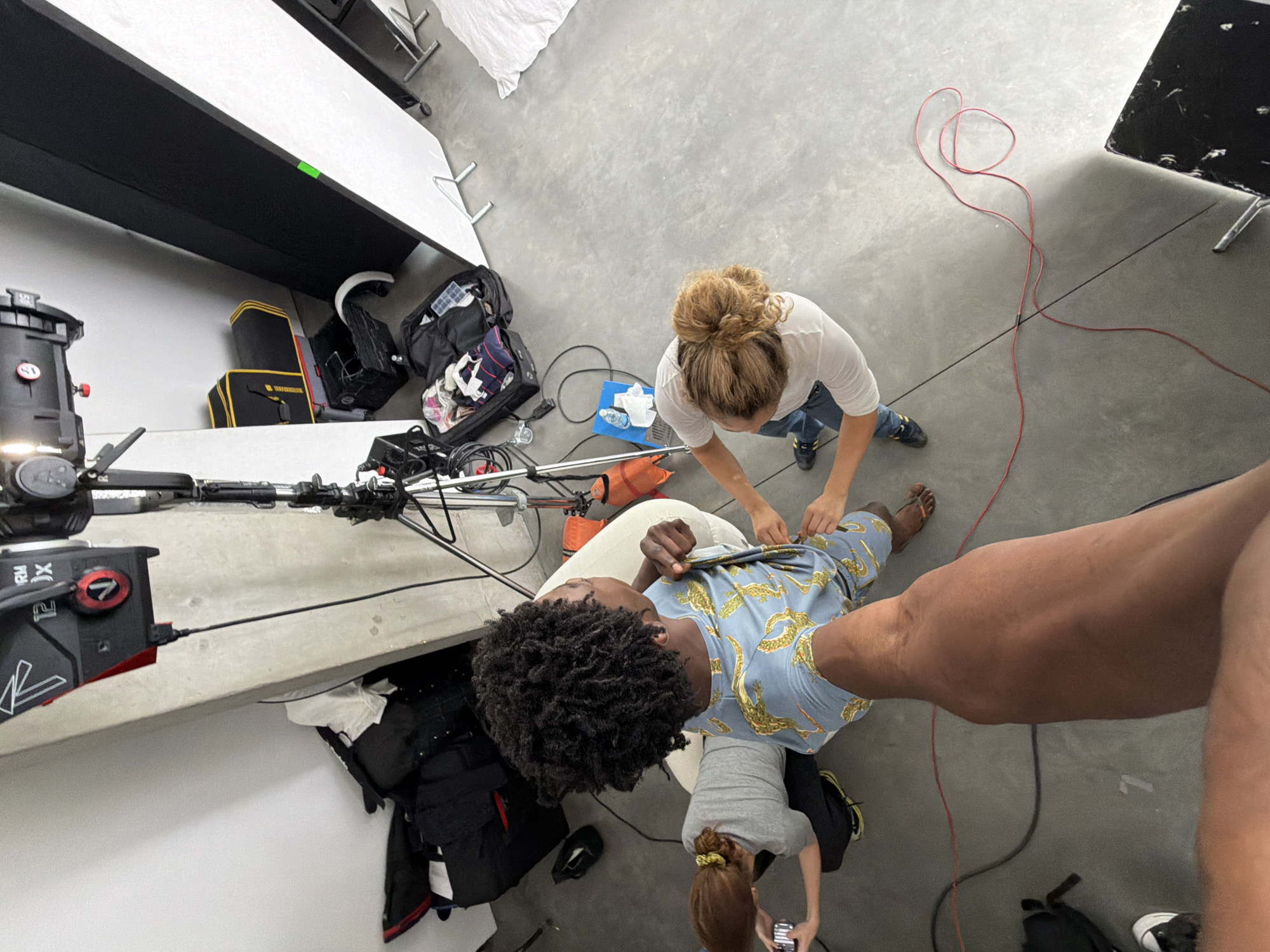 The image, shot from an elevated perspective, depicts an indoor scene, likely a photography or film studio. The location appears to be a large, open room with a smooth, light-grey concrete floor. Several individuals are present: one person is sitting in a chair wearing a blue shirt patterned with yellow alligators. A woman with her hair in a bun is standing near the seated person, appearing to adjust their clothing. Another person, with their hair in a bun is holding a phone. A large arm is also present in the image on the right. Studio equipment is visible, including a camera, lighting, and what appear to be stands or platforms. Bags and cases, possibly containing equipment, are scattered throughout the space. Red wires are on the floor. The image does not appear to be in London.