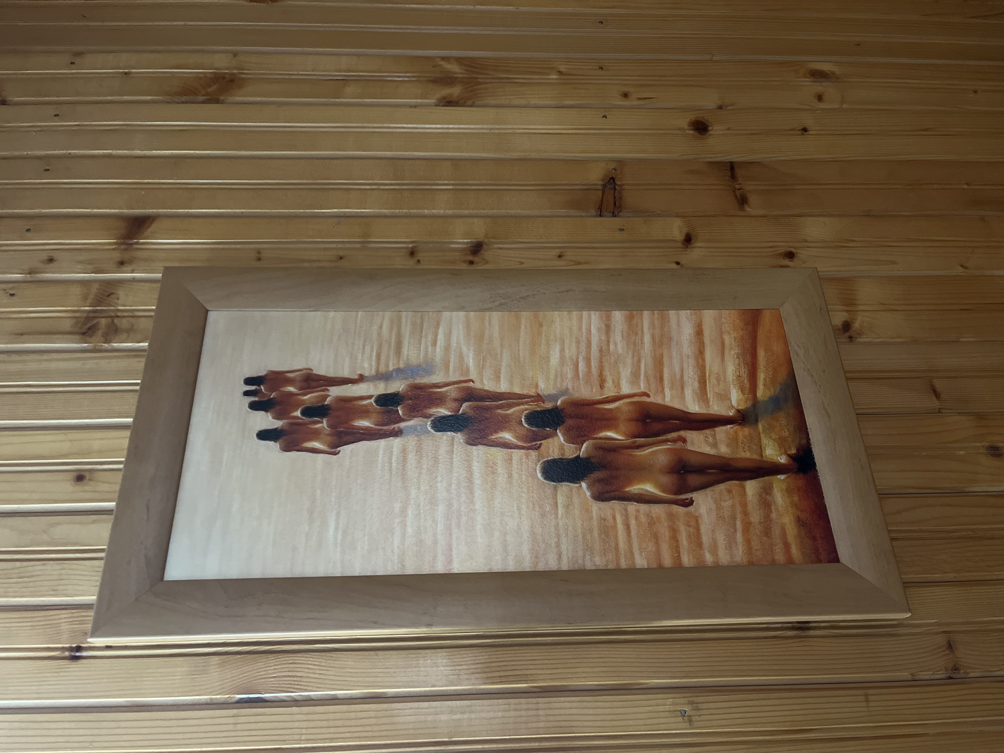 A framed painting of a line of women walking away from the viewer on a sandy surface hangs on a wood-paneled wall. The painting is oriented vertically and is framed with a light-colored wood frame. The women are depicted from behind, and their backs and buttocks are visible. They are all walking in the same direction, forming a diagonal line from the bottom right to the top left of the painting. The lighting in the painting appears to be warm, suggesting a sunset or sunrise. The wood-paneled wall is made of vertical planks of light-colored wood with visible knots and grain. The painting is positioned on the wall such that the top of the frame is roughly level with the viewer's eye.