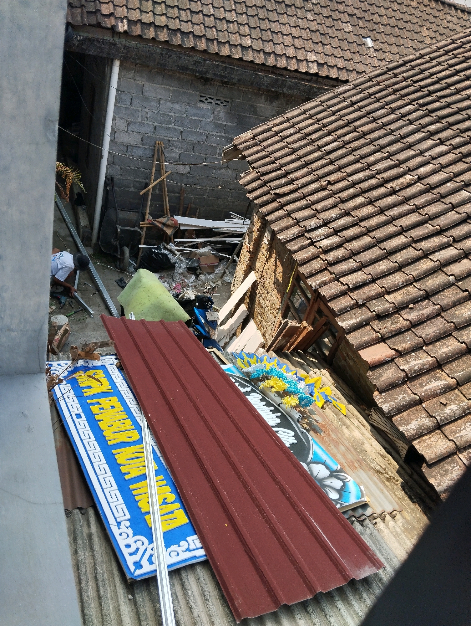 The image depicts a scene of what appears to be construction or demolition activity in a residential area in Salatiga, Indonesia. The foreground is dominated by a reddish-brown corrugated metal roof, likely newly installed or salvaged. To the left, a blue and white sign with yellow lettering is visible, suggesting it might be a directional sign or part of a business. The lettering on the sign, though partially obscured, reads "PEMBURUKUIAI YA".
Behind the sign, construction materials such as metal beams and what looks like a green tarpaulin are scattered on the ground, along with various debris, including discarded plastic, boxes, and a black trash bag. A person wearing a white t-shirt and a dark cap is visible in the mid-ground, appearing to be working with a drill on a metal structure. Next to them, there is a wheelbarrow and a motorcycle.
The background reveals a rustic brick wall and a tiled roof of an older building. The overall environment appears to be an outdoor space, possibly a backyard or an alleyway, characterized by a mix of old and new building materials and an ongoing activity. The lighting suggests it is daytime, with shadows cast by the structures.