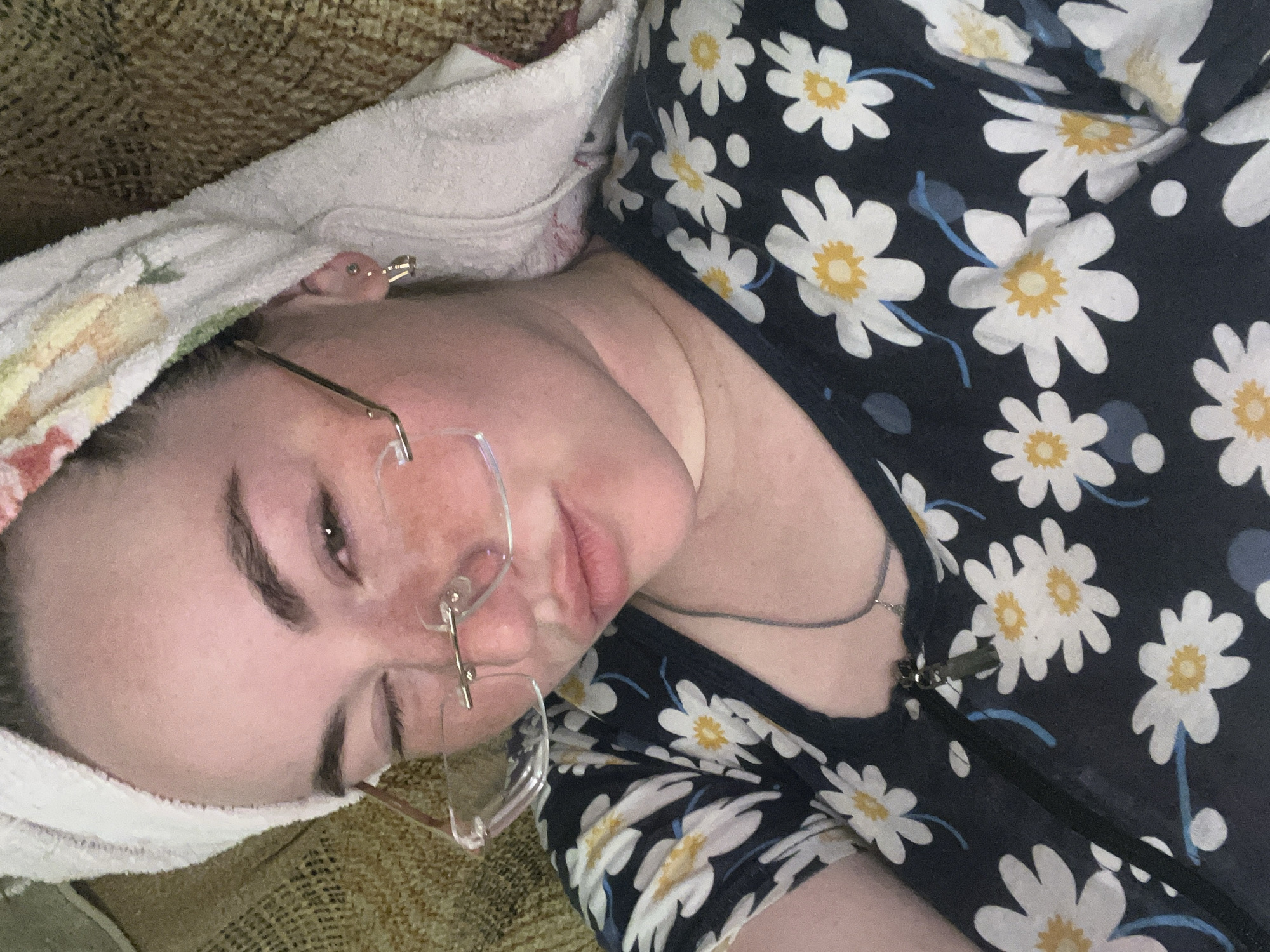 A woman with fair skin is lying on a textured tan surface, possibly a couch or bed, with her head resting on a white towel that has some floral patterns. She is wearing rimless glasses. The woman is wearing a dark-colored top with a daisy floral pattern, and what appears to be a zipper pull is visible near her chest. A necklace is partially visible on her chest as well. The location in the city of Сосново, Russia, cannot be determined from the visual information provided.
