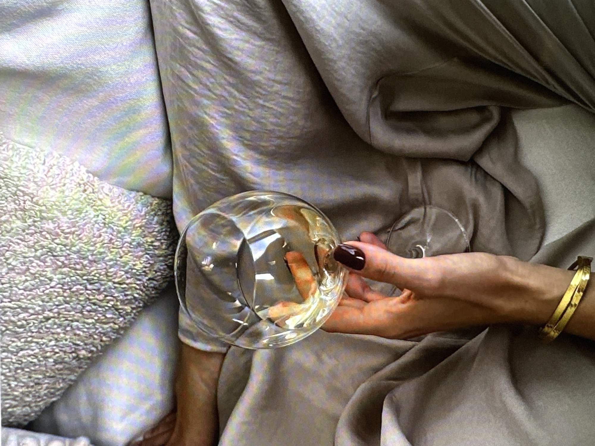 A top-down shot shows a woman holding an empty wine glass in her right hand. She is wearing a gold bracelet on her right wrist and has dark polished fingernails. Her hand and forearm are resting on what appears to be a soft, draped fabric in varying shades of grey. To the left of the wine glass is a textured cushion. The scene seems to be indoors. There is no information about the location being in Yerevan, Armenia.