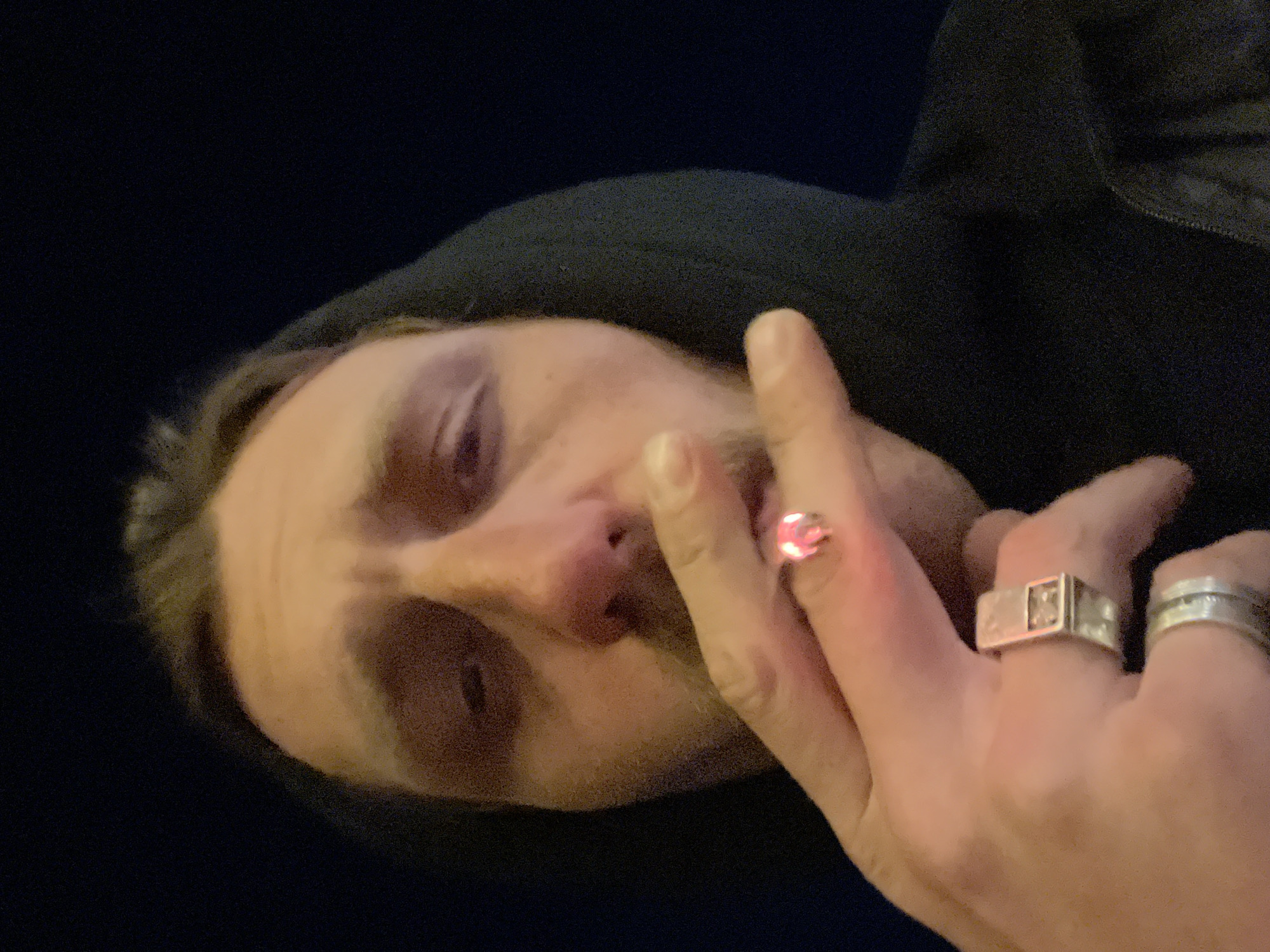 A low-angle close-up shot of a person's face and hand, positioned horizontally. The person is wearing a black hooded garment. Their face is pale, with reddish eyes and a prominent nose. They have a mustache. The hand is in the foreground, adorned with multiple rings: one on the pinky finger with a round stone, and two rings on the ring finger, one a wide band with a square or rectangular design. The background is a dark, undefined space. The location is unspecified.