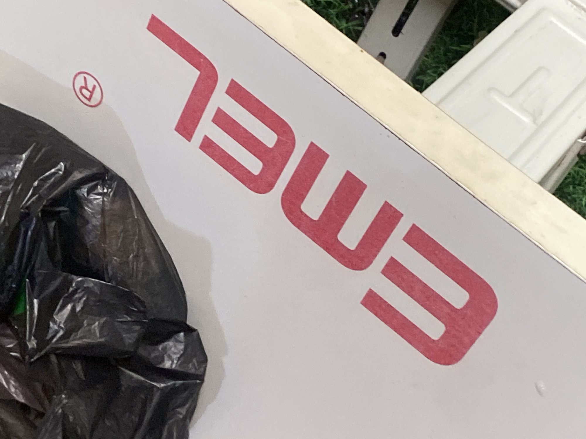 A close-up, high-angle shot shows a black plastic bag and the red text "EMEL" on a white surface. To the left of the text is a red circle with an "R" inside, indicating a registered trademark. The white surface appears to be part of a table or desk. In the background, a portion of a white metal object, possibly a sewing machine, is visible, resting on a green surface that resembles grass. The scene is likely indoors, given the lighting. The location is described as Jauro Yinu, Nigeria.