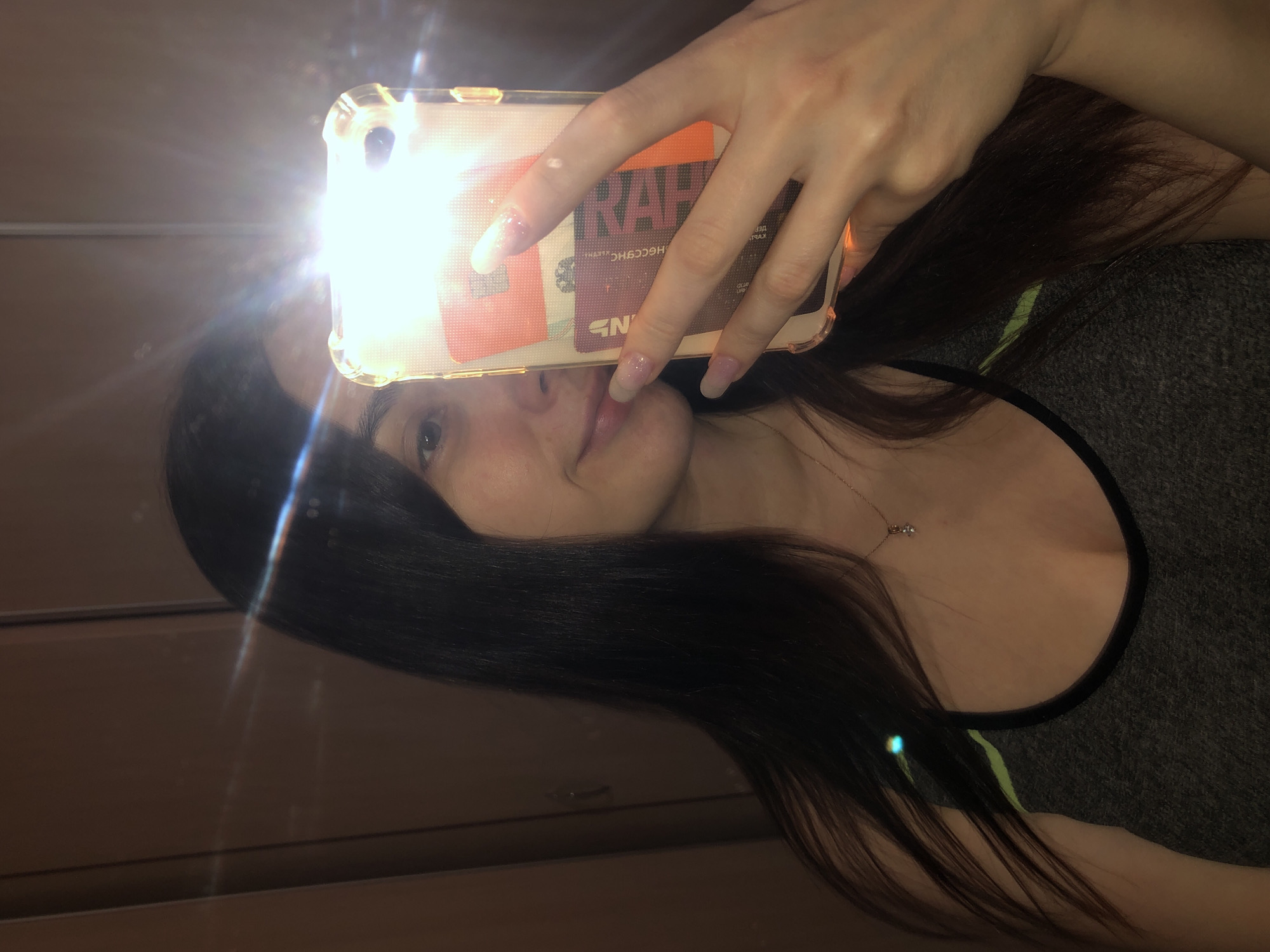 A young woman is taking a selfie in what appears to be an indoor location. She is holding a phone with a clear case, which contains cards. The bright flash from the phone obscures parts of her face. She has long, dark hair and is wearing a gray tank top with black trim. A delicate necklace is visible. The background appears to be a dark surface, possibly a wooden wall or cabinet. The location is not discernible as the photo lacks specific identifiers to associate it with Noviy, Russia, or any particular place.