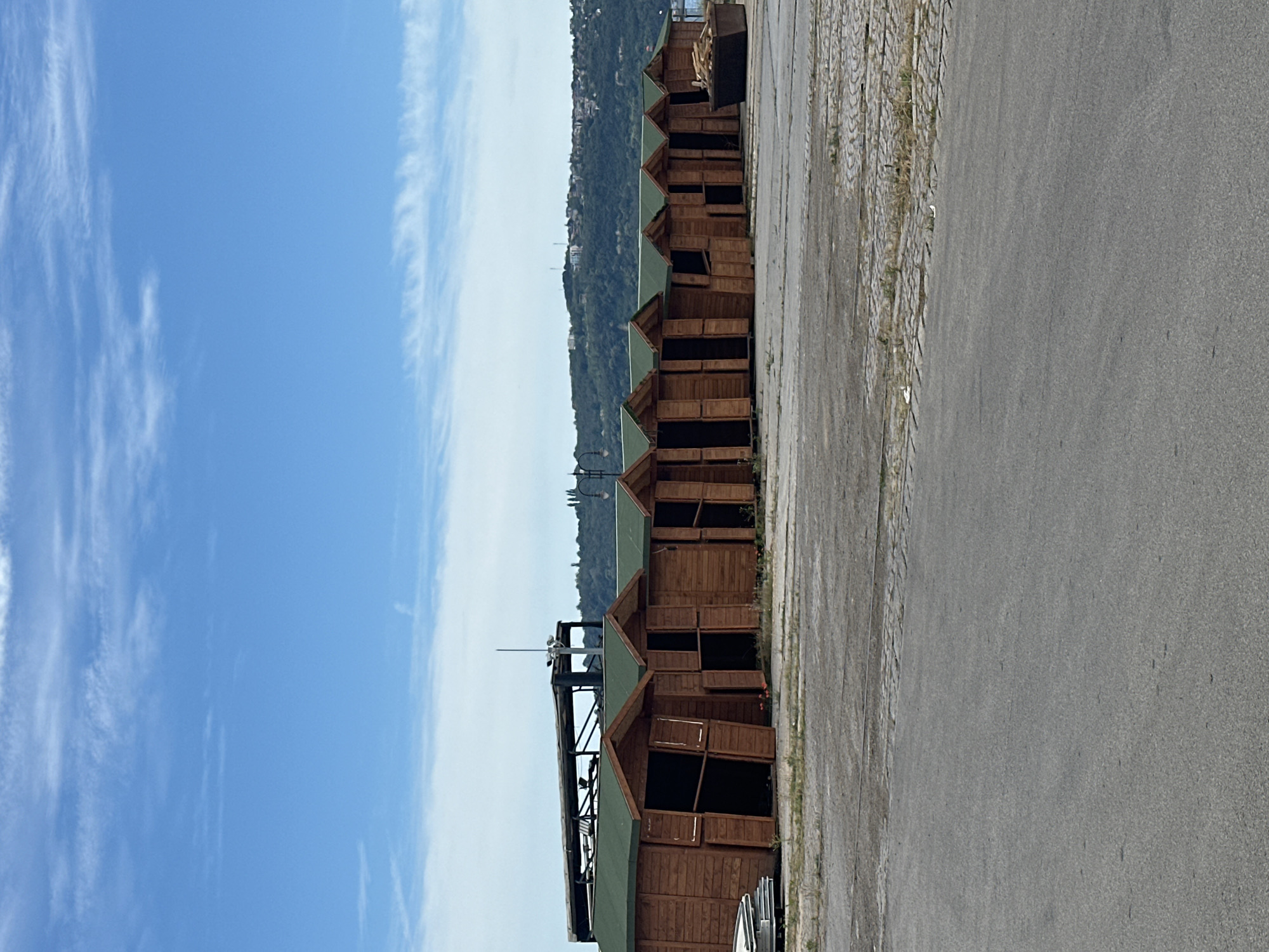 A long line of identical wooden market stalls with green roofs are arranged on an asphalt surface. The stalls are open, revealing dark interiors. In the background, a hill covered in trees and buildings slopes down towards a body of water. The sky is a bright blue with scattered white clouds. A large dumpster filled with wood is visible to the right of the stalls. The overall scene suggests an outdoor market area that is currently not in use.