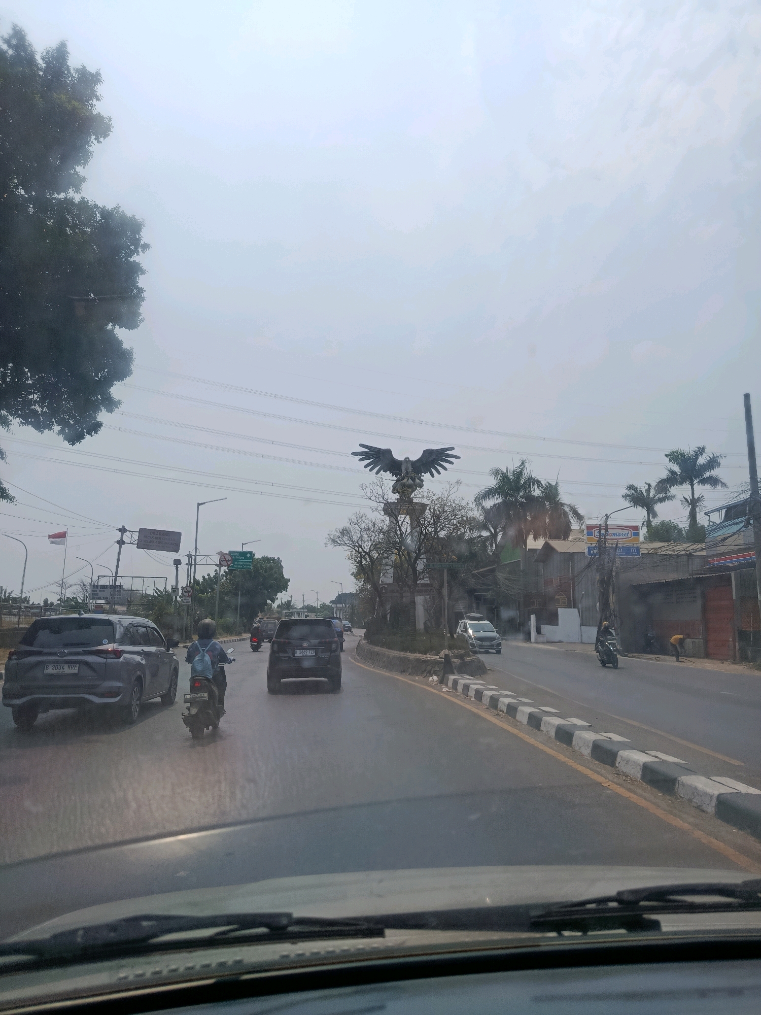The image depicts a street scene in East Jakarta, Indonesia, on a cloudy day. A four-lane road is visible, with vehicles traveling in both directions. On the left side of the road, a gray SUV with license plate B 2034 KRE is parked. A person on a motorcycle, wearing a blue backpack, is riding in the left lane. Further down the road, another dark SUV with license plate B 2509 TZO is in the right lane.
In the background, a large eagle statue is prominent, mounted on a pedestal. Palm trees and other vegetation are scattered throughout the scene. Buildings, including a store with a "Indomaret" sign, line the right side of the road. Power lines crisscross the sky. A red and white Indonesian flag is flying on a pole on the left side. The road is marked with yellow and white lines, and a concrete curb with black and white stripes separates the lanes. A person is visible in the distance on the right side of the road, appearing to be working. The overall impression is of a typical urban street environment in Indonesia.