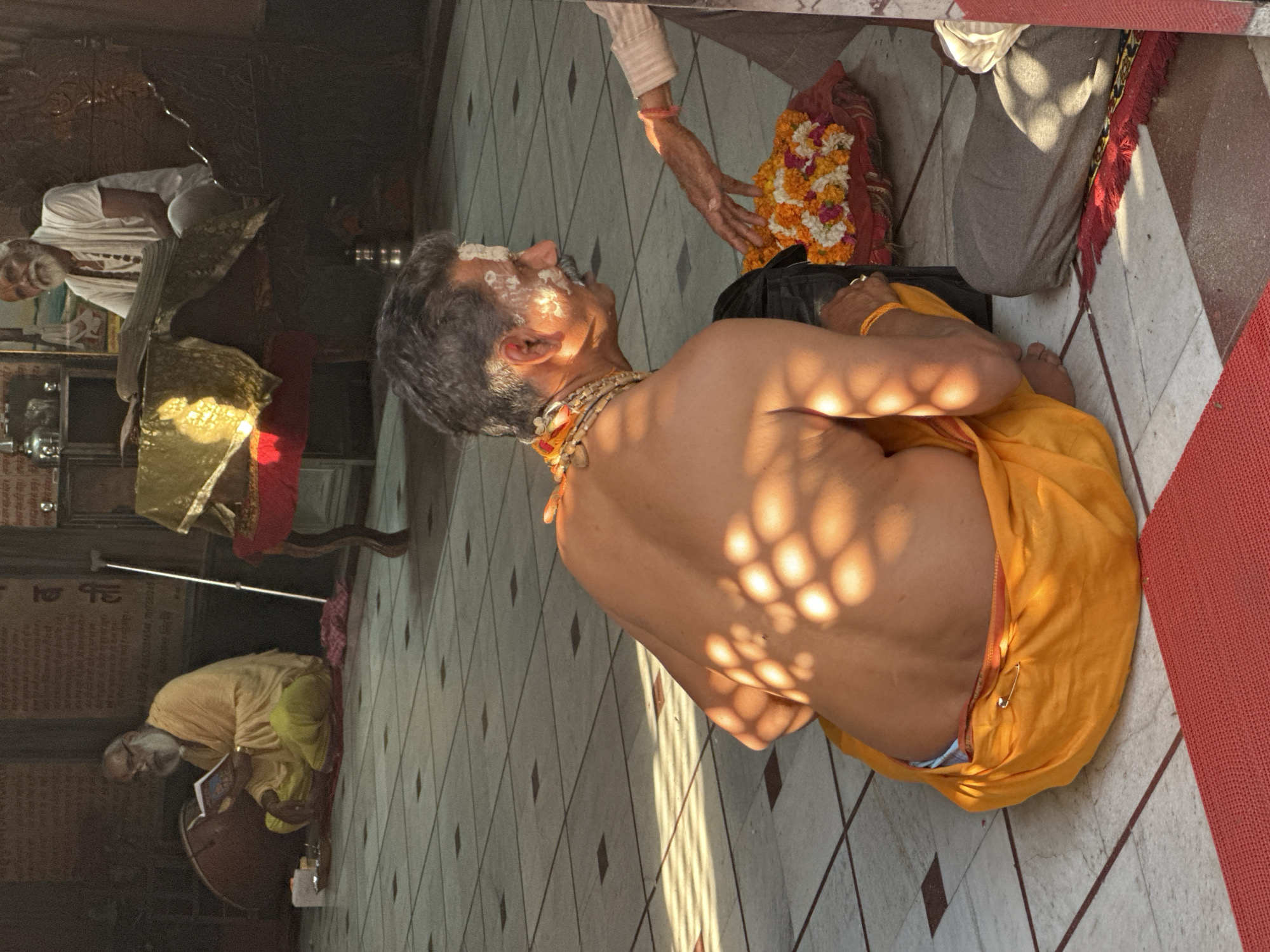 The image shows a man with white markings on his face and wearing a beaded necklace, seated in a temple. He is shirtless and draped in an orange garment. Sunlight filters through the space, casting patterned shadows on his back. Another man stands beside him, holding a garland of flowers, possibly performing a ritual. In the background, a seated older man is playing a drum. The scene is taking place in Vrindavan, India.
