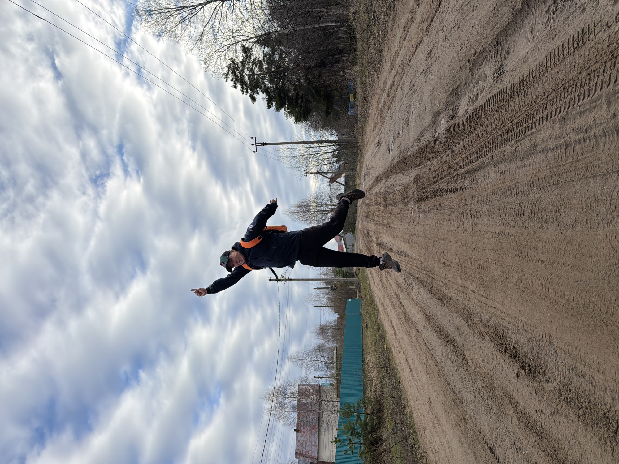 The image, taken in Chaikovsky, Russia, features a person captured mid-air, appearing to jump or leap horizontally across a rural dirt road. The photograph is rotated 90 degrees counter-clockwise, creating an illusion of the individual floating parallel to the ground. The person, an adult, is dressed in a dark jacket with an orange strap (possibly part of a backpack), dark pants, athletic shoes, and a dark baseball cap. Their arms are extended, with one arm pointing forward, and their legs are also outstretched, suggesting a dynamic, playful action.
The scene depicts a rustic environment with a wide dirt road dominating the foreground and middle ground (when oriented correctly). The road is marked with numerous tire tracks. To the left, a teal corrugated metal fence partially hides small, simple buildings or structures, with bare deciduous trees and some smaller evergreens behind them. On the right, a denser line of trees, including evergreens and leafless deciduous trees, borders the road. Utility poles and power lines crisscross the upper portion of the frame.
The sky is bright with large, prominent white clouds and visible patches of blue, indicating daylight, likely late morning or afternoon. The bare branches of the deciduous trees suggest a cool season, such as early spring or late autumn. No visible text is present in the image. The overall impression is one of energetic activity within a natural, semi-rural setting.