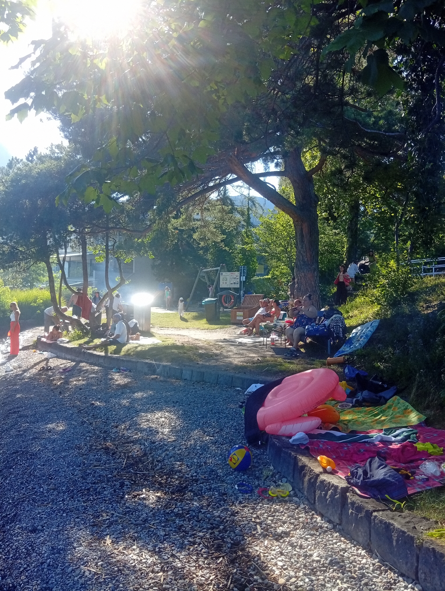 The image depicts a sunny day scene at a lakeshore, likely in Hergiswil (NW), Switzerland, given the context. The foreground is dominated by a pebbled beach with scattered debris and shadows from overhead trees. A stone embankment runs along the edge of the beach, on top of which several colorful blankets, towels, and inflatable toys are spread out. Among the inflatables is a large pink ring float and an orange float. A striped beach ball and a pair of goggles are also visible on the pebbles.
Moving further into the scene, a group of people are gathered under the shade of large trees. Some are seated on chairs and blankets, appearing to be engaged in conversation or relaxation. One individual, a woman with blonde hair, stands near the water's edge on the left, wearing a white top and an orange skirt. Further back, a group of people are seated around a table and some are standing. In the background, there's a glimpse of buildings, possibly houses or facilities, and more lush greenery. A playground structure with a swing set is visible to the right of the central gathering area, and a signpost with multiple information boards stands nearby. The overall atmosphere suggests a casual outing by the lake, possibly a picnic or a day at the beach. The bright sunlight creates strong highlights and shadows throughout the scene, indicating a clear and warm day.