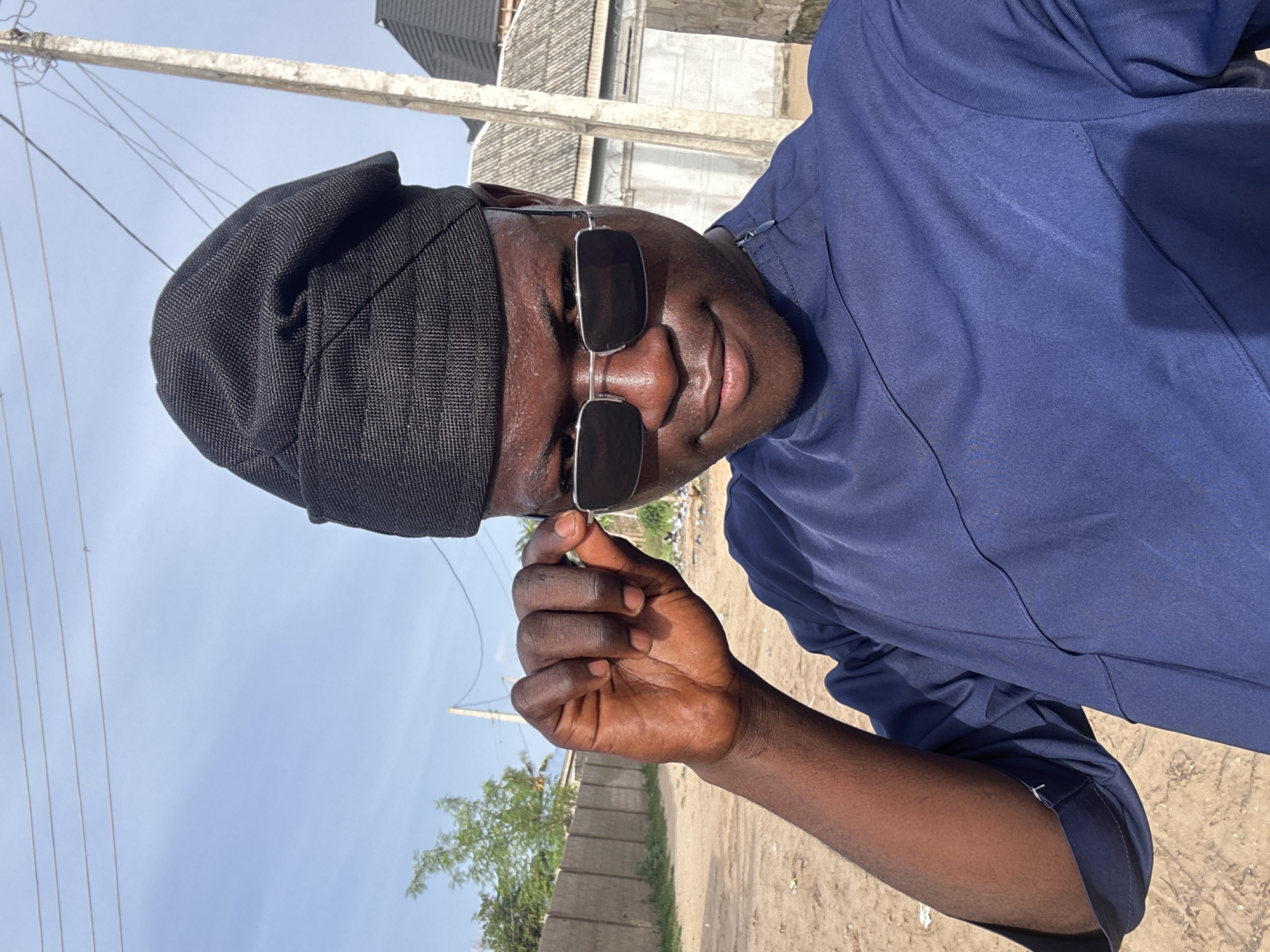 An eye-level photograph depicts a dark-skinned man with a shiny complexion, wearing a black cap and dark sunglasses. He is dressed in a blue shirt or robe. His right hand is raised, appearing to adjust his sunglasses. The man is located outdoors in a sunny environment in the city of Badagry, Nigeria. The background includes a dirt ground, a cement or concrete fence, trees, power lines, and buildings in the distance. The image has been rotated counter-clockwise.