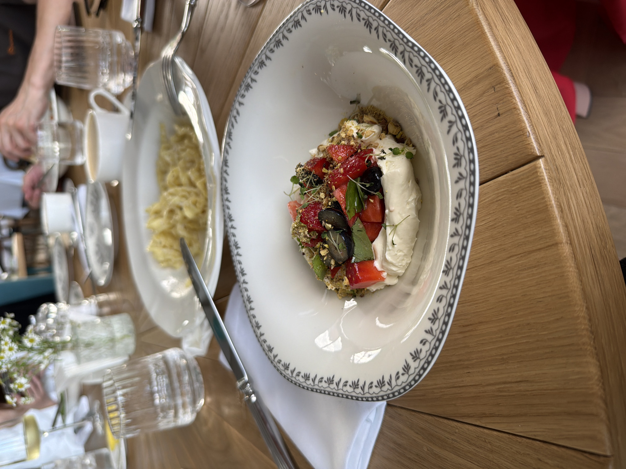 This is a close-up, overhead shot of a meal on a wooden table. In the foreground, a white bowl with a decorative rim holds a dessert. The dessert appears to be a mix of berries, including strawberries and blueberries, crumbled nuts, and a dollop of whipped cream or mascarpone cheese. Small green herbs are sprinkled on top. To the left of the dessert bowl, a plate holds a portion of pasta, possibly fettuccine, mixed with a light sauce. A fork rests on the pasta plate, and a knife is positioned on a white napkin below the dessert bowl. In the background, a person's hands are visible, reaching for a glass. Several clear glasses, some containing liquids and ice, are scattered on the table, along with a white cup and saucer. A small bouquet of white flowers with green stems is also visible to the left. The lighting suggests it could be daytime. The overall impression is of a restaurant setting, with a focus on the food and dining experience.