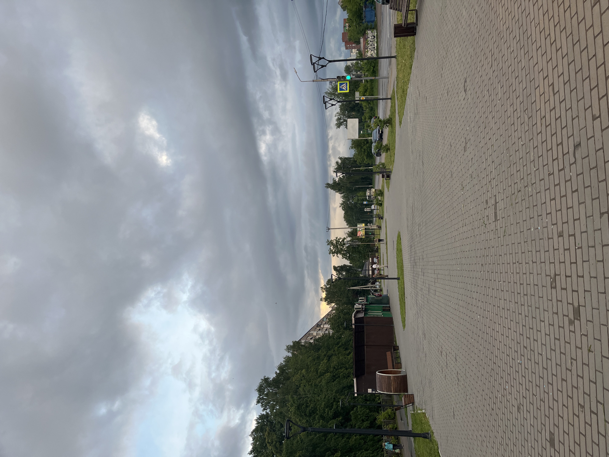 This image, oriented correctly, presents an outdoor scene in Chaikovsky, Russia, featuring a paved walkway bordering a street or park area under a heavily overcast sky.
In the foreground and midground on the right, a wide walkway composed of grey rectangular paving stones stretches into the distance. Along its left edge runs a strip of green grass, beyond which are trees and what appears to be a road or a parking area. Several dark-colored streetlights, wooden benches, and a large circular wooden planter or waste bin are spaced along the paved path.
On the left side of the scene, a dense line of green trees and bushes forms a natural barrier, partially obscuring background elements. Above the tree line, a traffic light displaying a green signal and a yellow pedestrian crossing sign are visible on utility poles. Further in the background, glimpses of buildings, possibly residential or commercial structures, can be seen beyond the foliage. A few indistinct vehicles are also visible on the road.
The upper portion of the image is dominated by a dramatic, overcast sky filled with elongated, layered grey clouds. Patches of brighter, lighter sky are visible where the cloud cover is thinner, suggesting some light is still penetrating, particularly towards the horizon on the left, hinting at a late afternoon or early evening time. A few blurry figures of people are faintly visible in the far distance along the walkway, engaged in no specific discernible activity. No legible text is visible within the image.