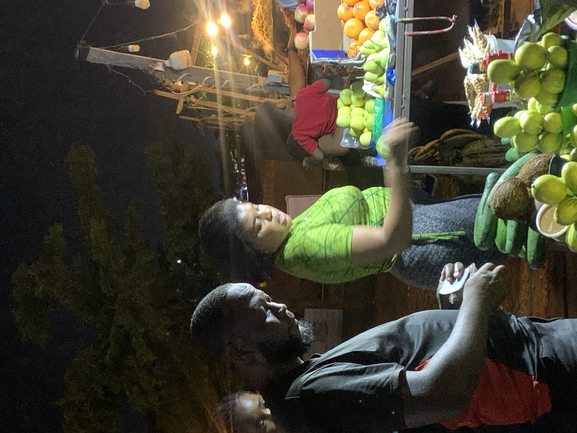 This is a nighttime, medium-shot photograph taken at a fruit stall in Galadimawa Village, Nigeria. The scene is illuminated by artificial lights, casting a warm glow on the produce and people.
In the foreground, a man with a dark complexion and a close-cropped beard, wearing a black t-shirt with a red undershirt visible beneath, is in profile, looking down at something in his hands. His hands are blurred, suggesting movement. To his right, a woman with dark, curly hair, wearing a bright green t-shirt with a subtle pattern, is looking downwards, also with her hands in motion. She appears to be interacting with the fruit stall.
The fruit stall itself displays a variety of produce. Piles of green apples and pears are visible in green and blue containers. Further back, oranges and possibly other citrus fruits are arranged on a shelf. To the far right, a pile of what appears to be a crispy snack or dried fruit is presented in a container. Several cucumbers are stacked in the lower left quadrant of the image, along with two coconuts. In the background, a young child wearing a red t-shirt is seated, facing away from the camera. The stall is set up outdoors, with a dark, bushy tree visible in the upper left portion of the frame. Various strands of string lights hang overhead, adding to the ambiance of a nighttime market.