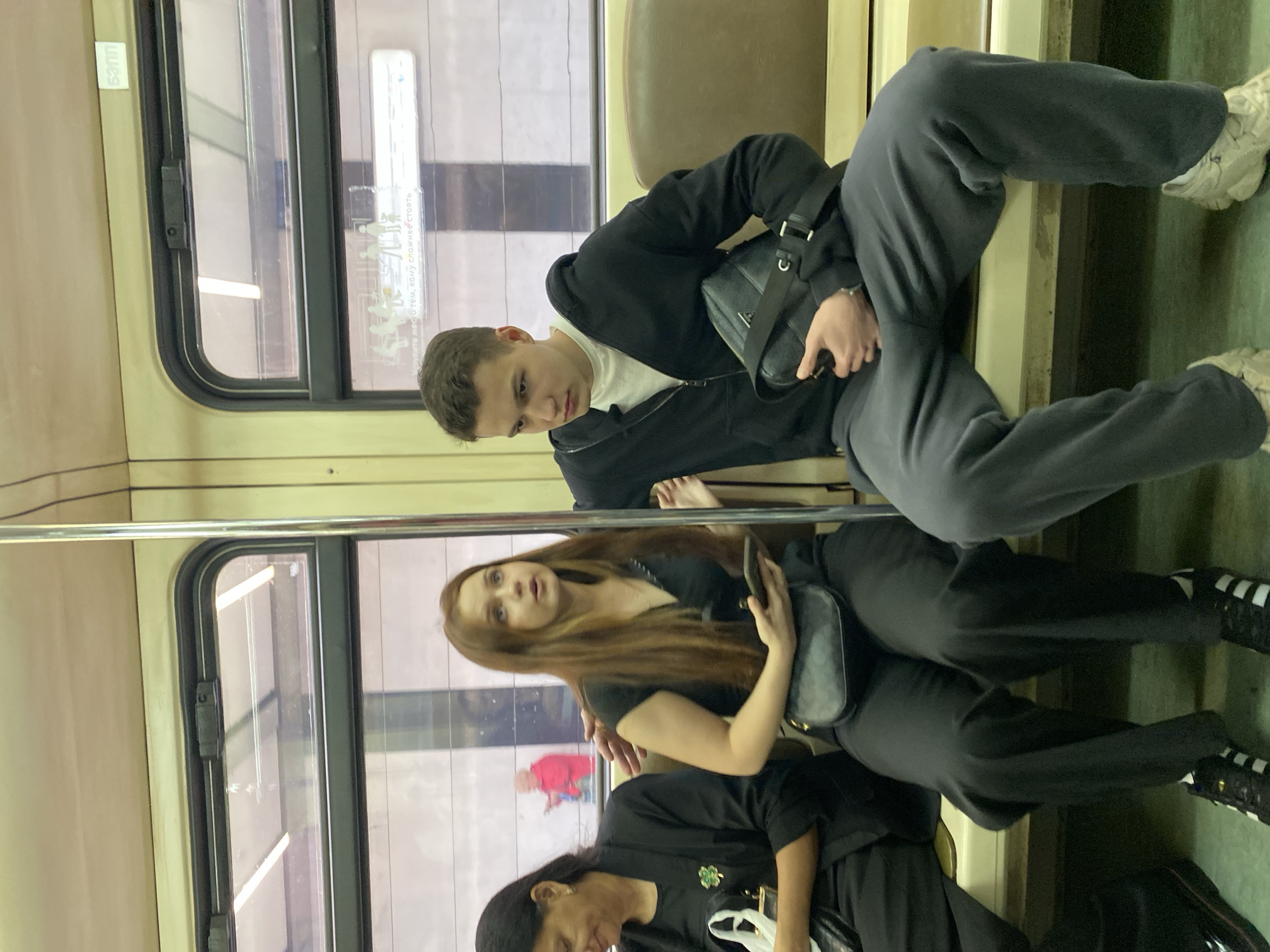 Inside a public transit vehicle, possibly a subway car, three individuals are seen. A young man in a black jacket and gray sweatpants sits with his legs spread out, occupying more than one seat. A woman with long brown hair is seated below him, appearing to look up in his direction while holding a mobile phone. A third person is partly visible below her, wearing a black top with a white object. The environment includes windows with blurry urban scenes visible outside, and a metal handrail is present. The overall setting suggests an urban environment in Unknown, Unknown.